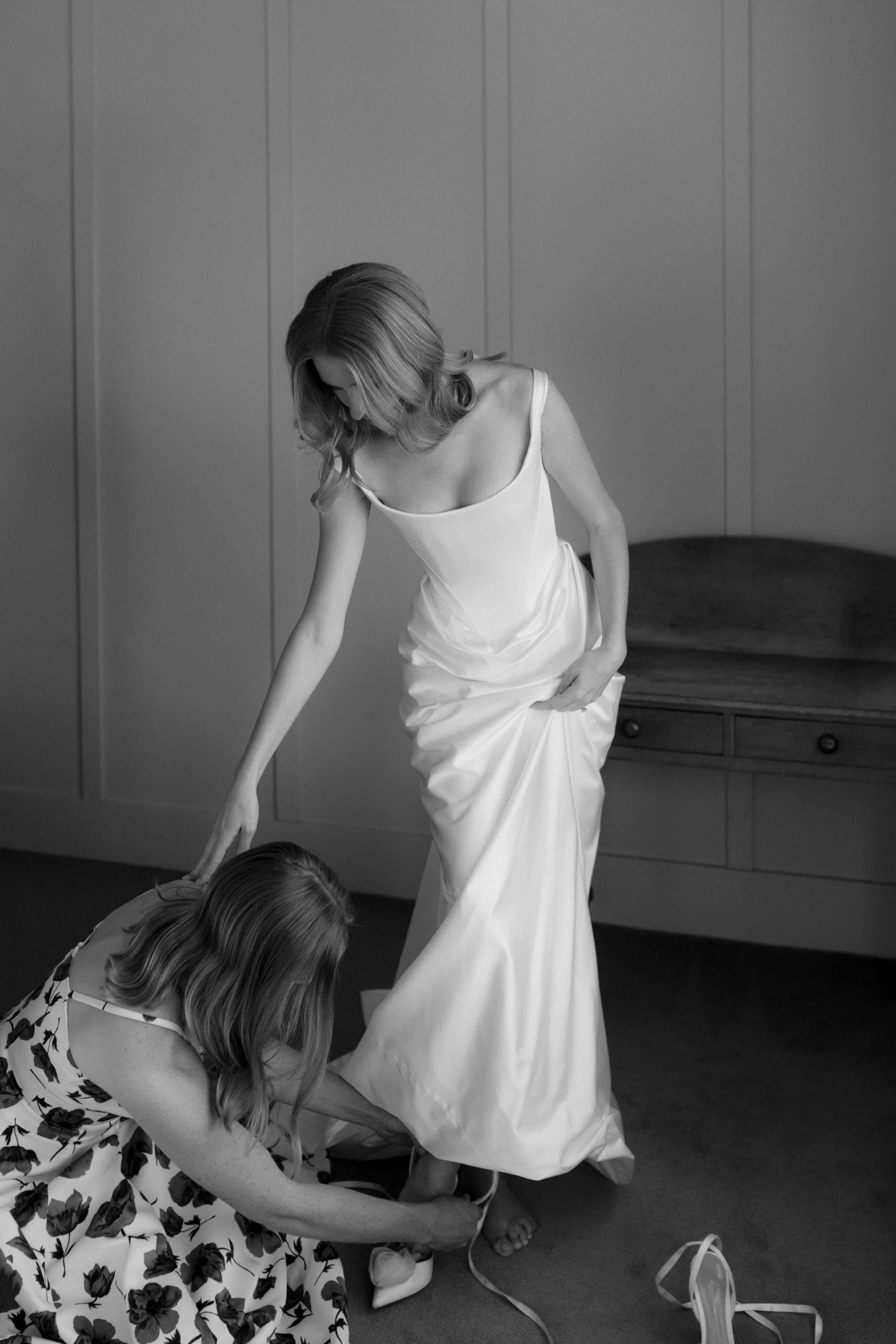 A woman in a white gown is helping another woman put on shoes in a bedroom.