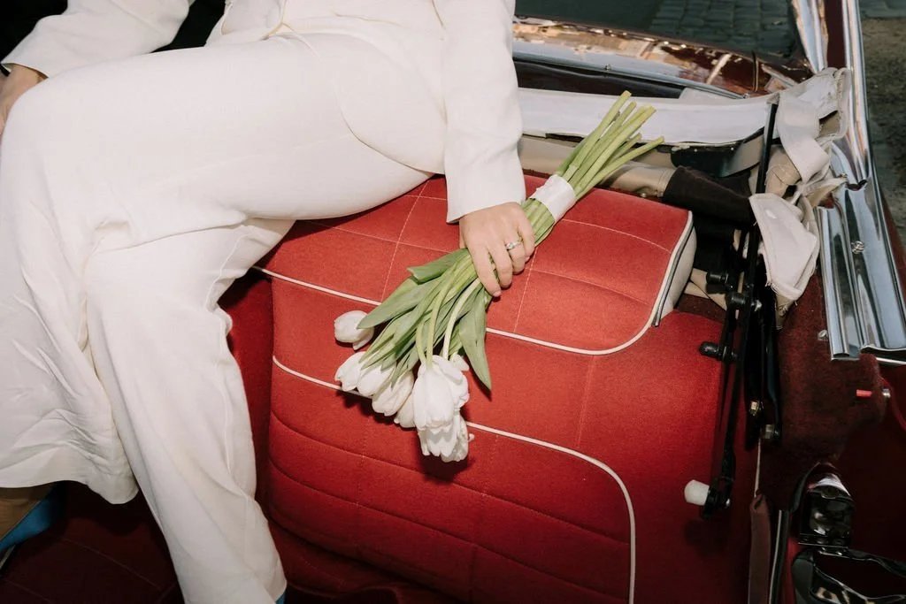Person dressed in white clothing sitting on a vintage red car seat, holding a bouquet of white tulips.