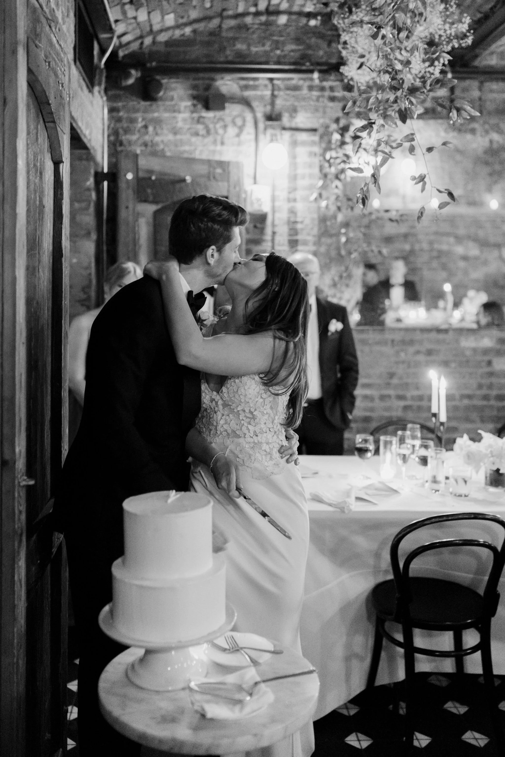 A black and white photo of a newlywed couple kissing at their wedding reception, with a wedding cake in the foreground and guests in the background.