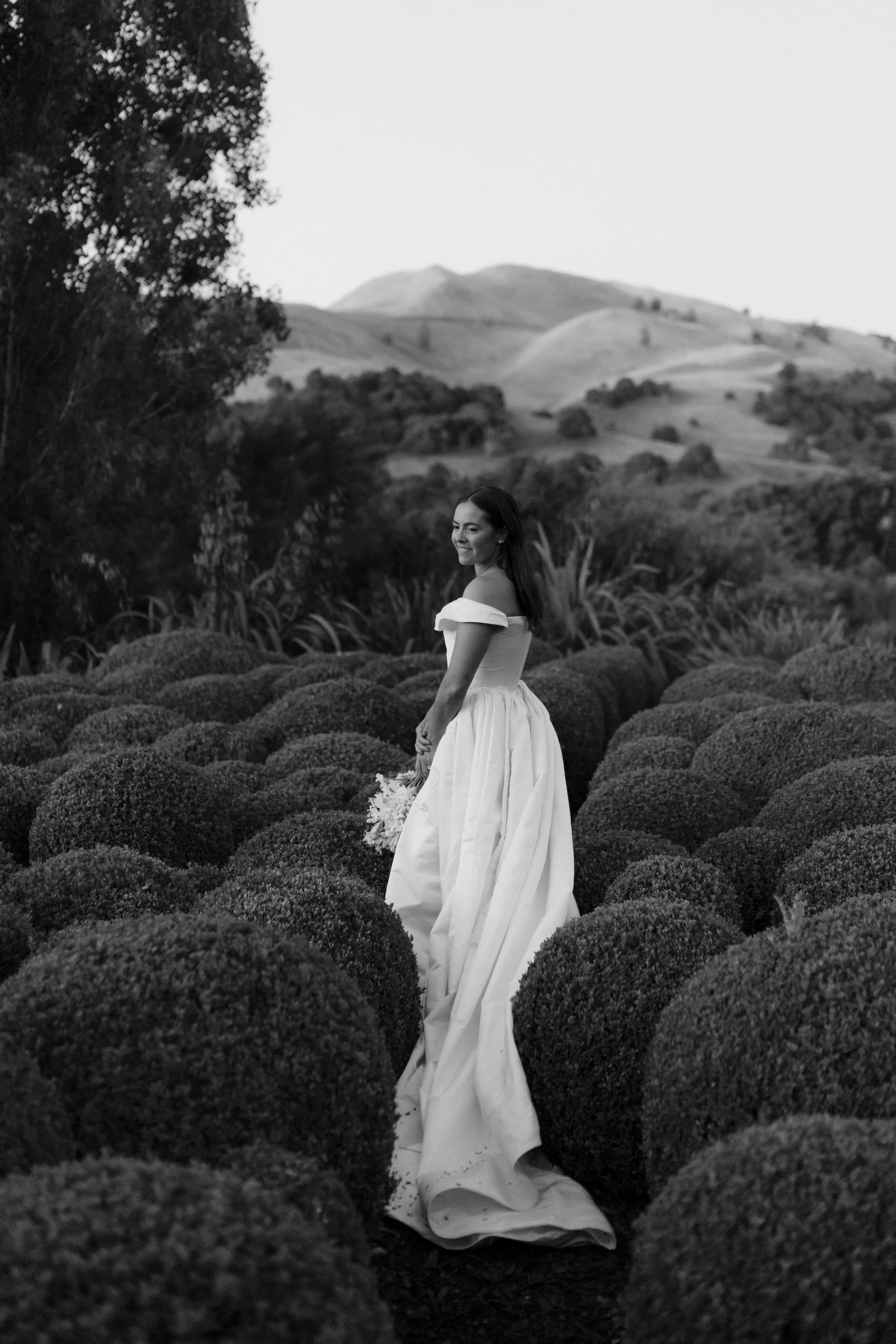 A woman in a wedding dress standing among bushes in a natural landscape, holding a bouquet, in black and white.