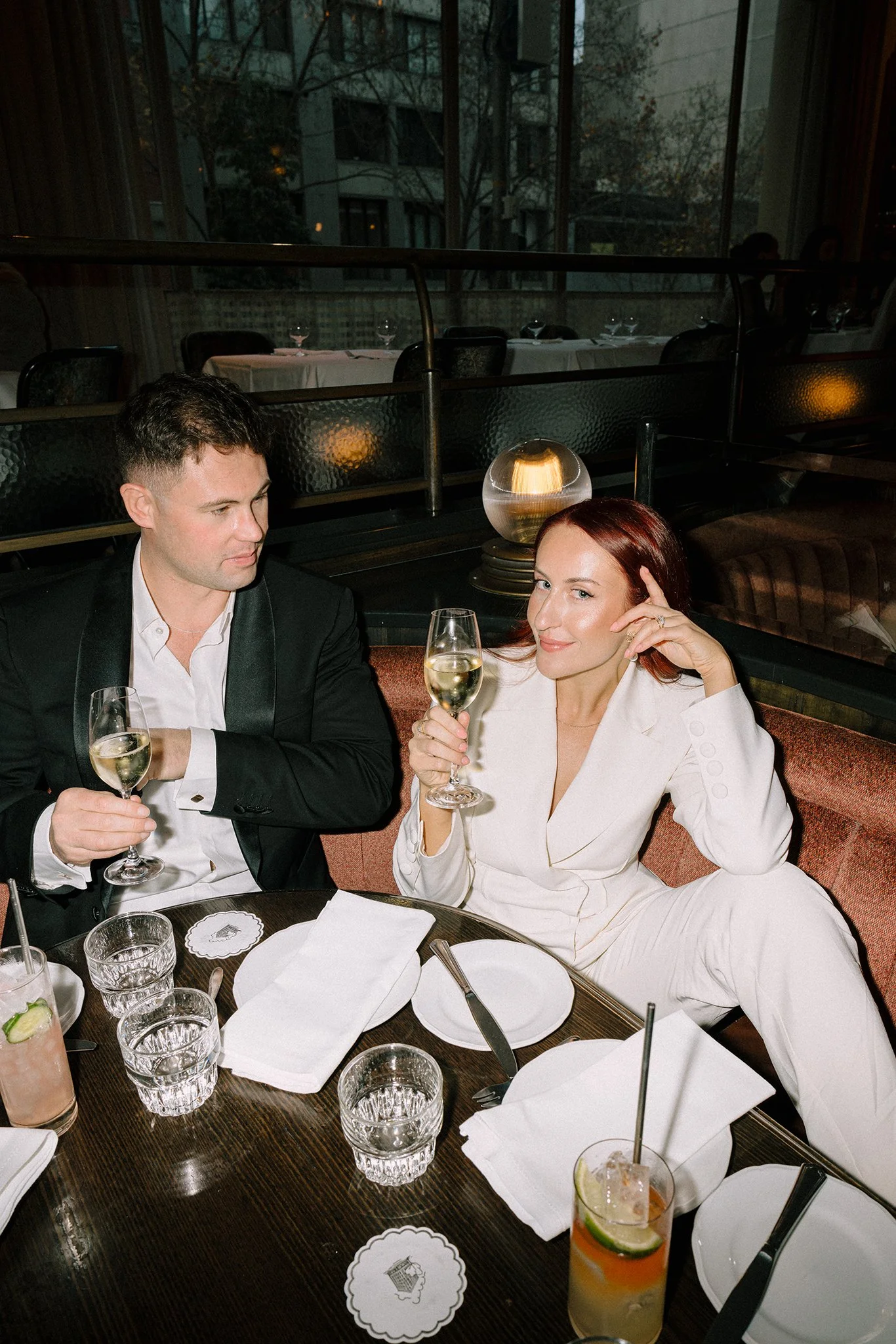 A man and woman sitting at a restaurant table, dressed formally, holding glasses of white wine, with various drinks and table settings visible.