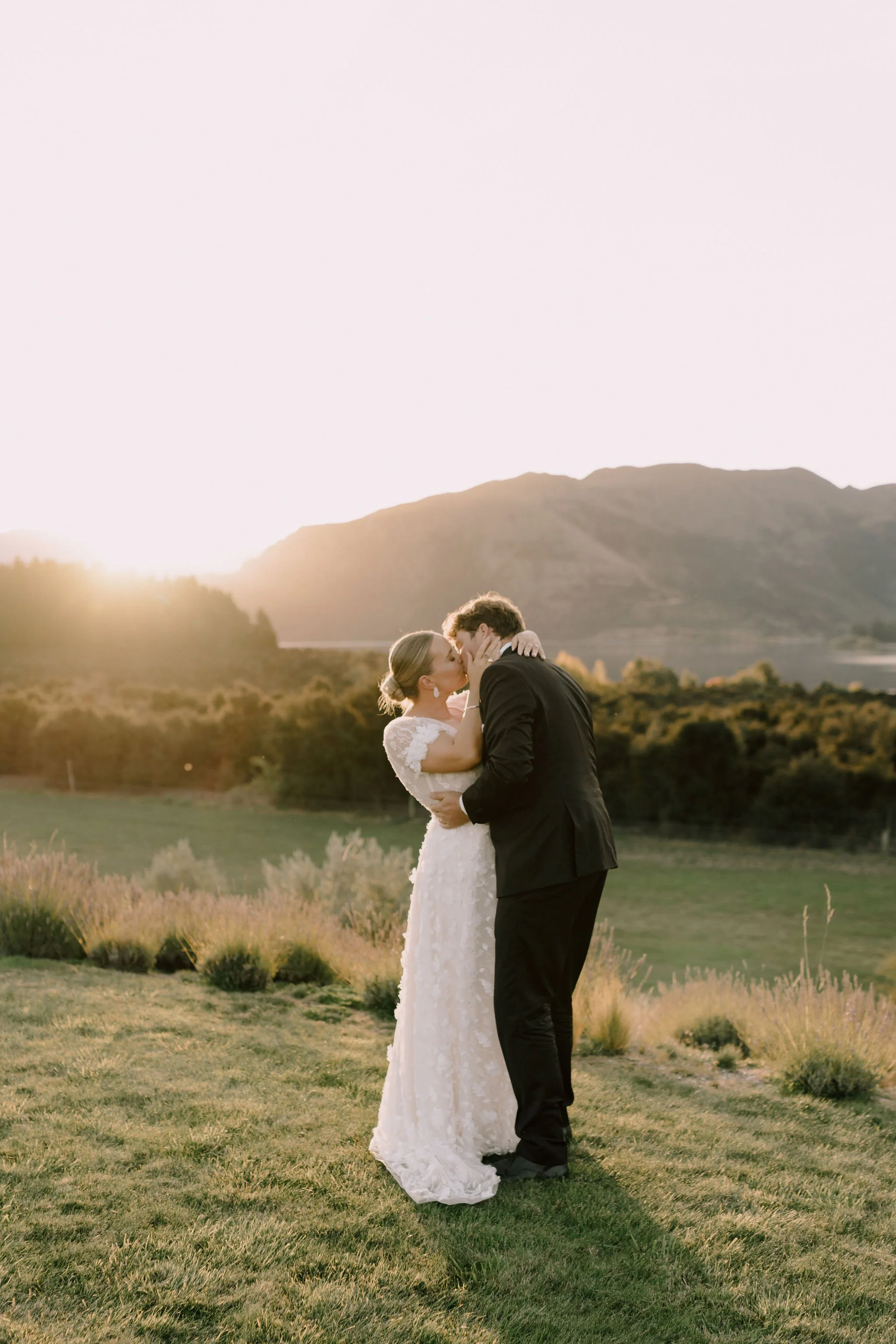 A bride and groom kissing outdoors at sunset, standing on green grass with mountains in the background.