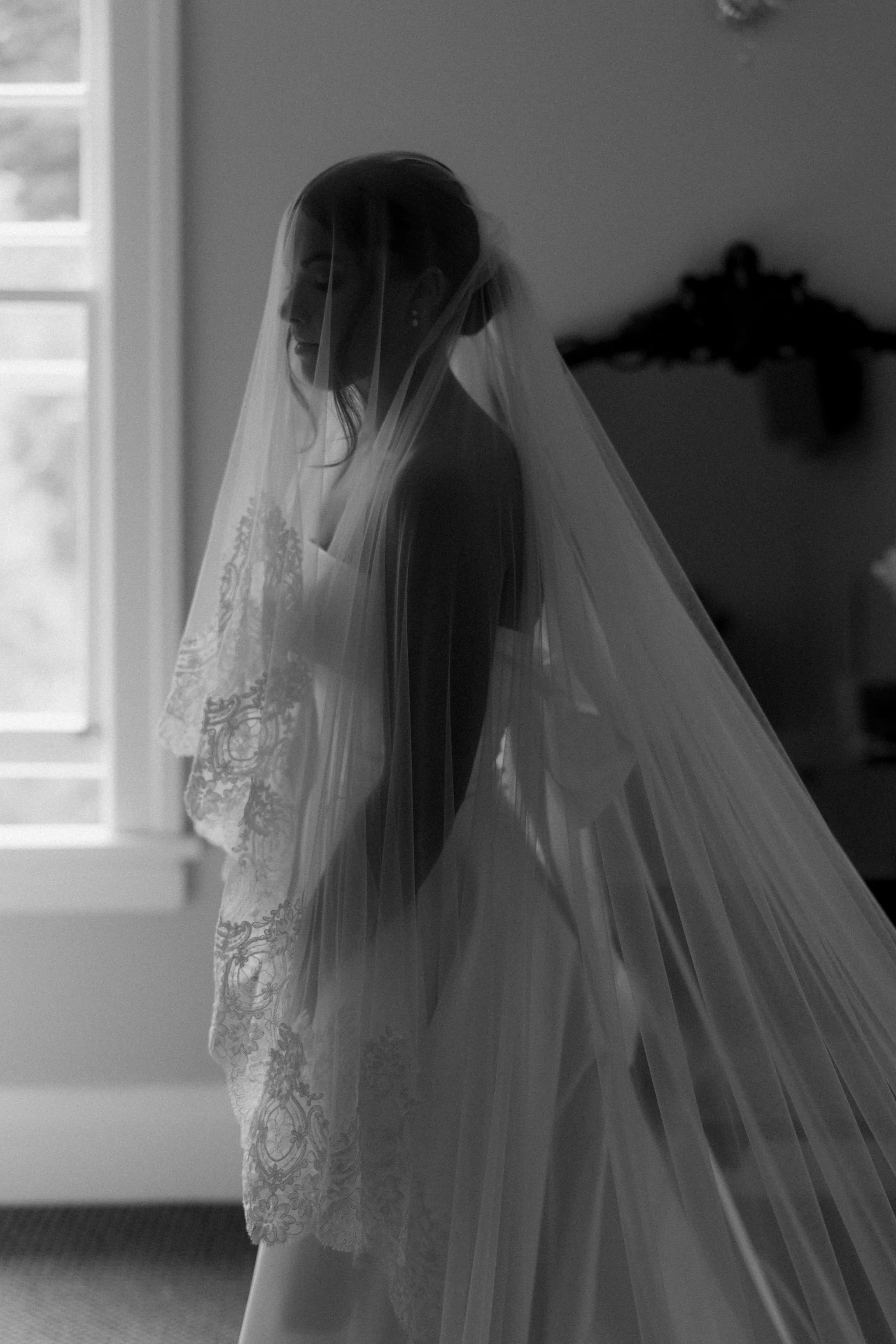 A bride in a wedding dress with a veil standing in a room near a window, with her face turned slightly downward.