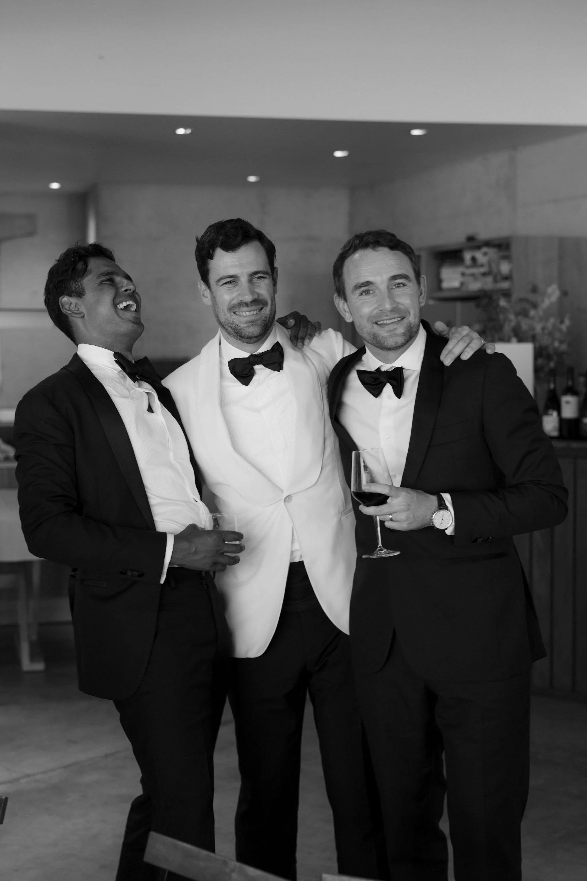 Three men in tuxedos and bow ties celebrating at a formal event, smiling and holding drinks, in a modern indoor setting.