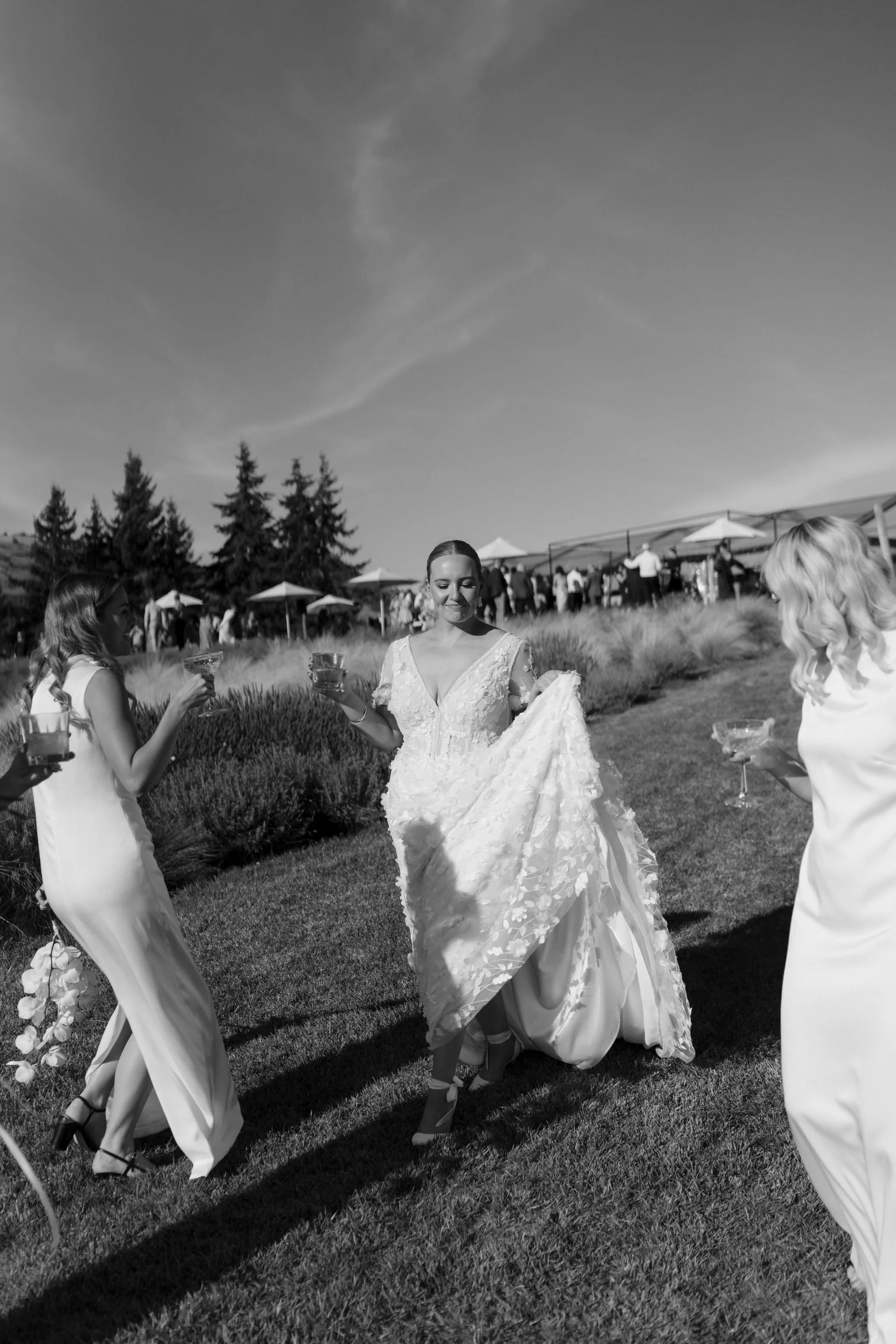 Bride in wedding dress holding gown train, surrounded by bridesmaids holding drinks, outdoor wedding reception setting with tents and guests