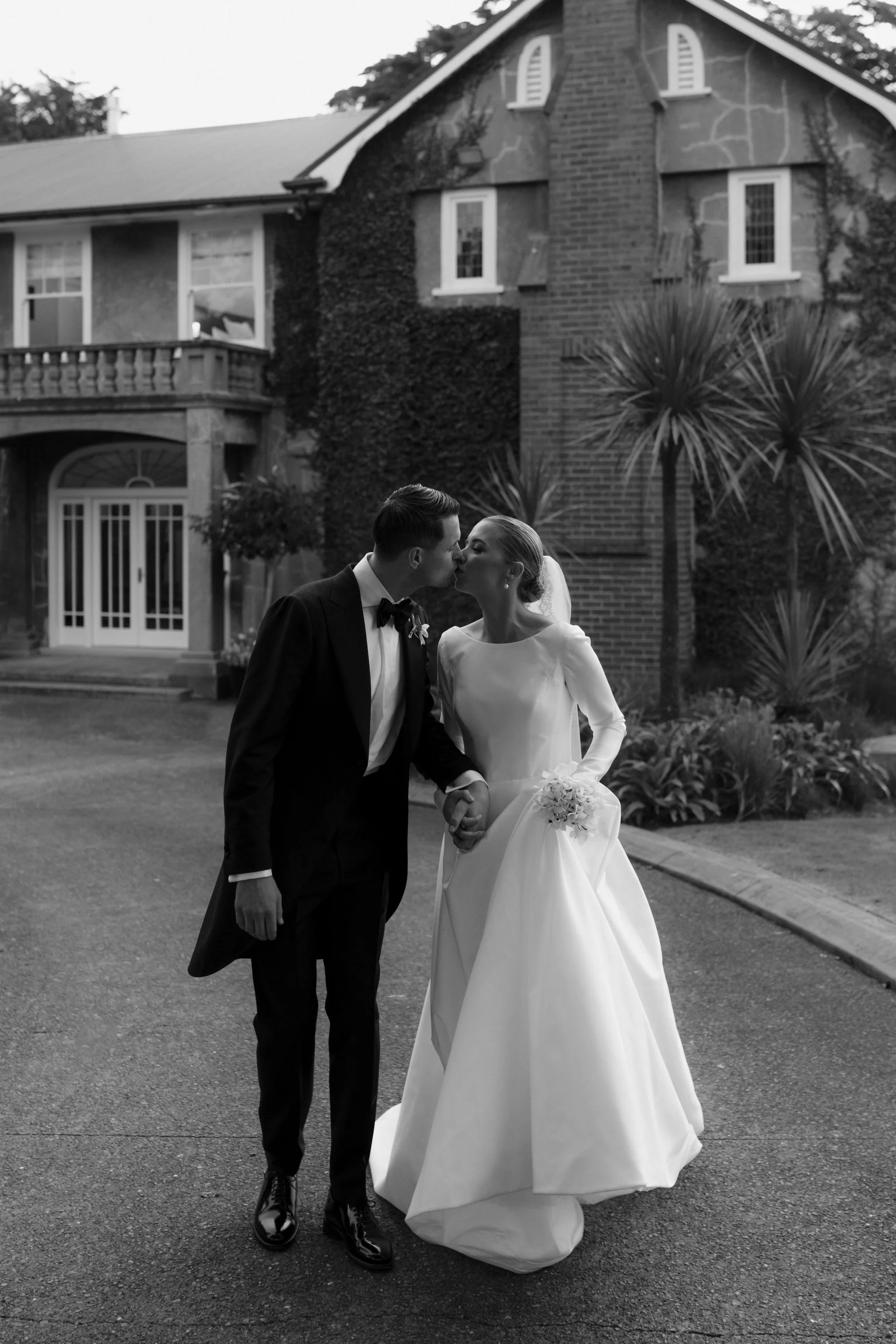 A black-and-white photo of a bride and groom holding hands and sharing a kiss outside a house, with the groom wearing a tuxedo and the bride in a wedding gown holding a bouquet.