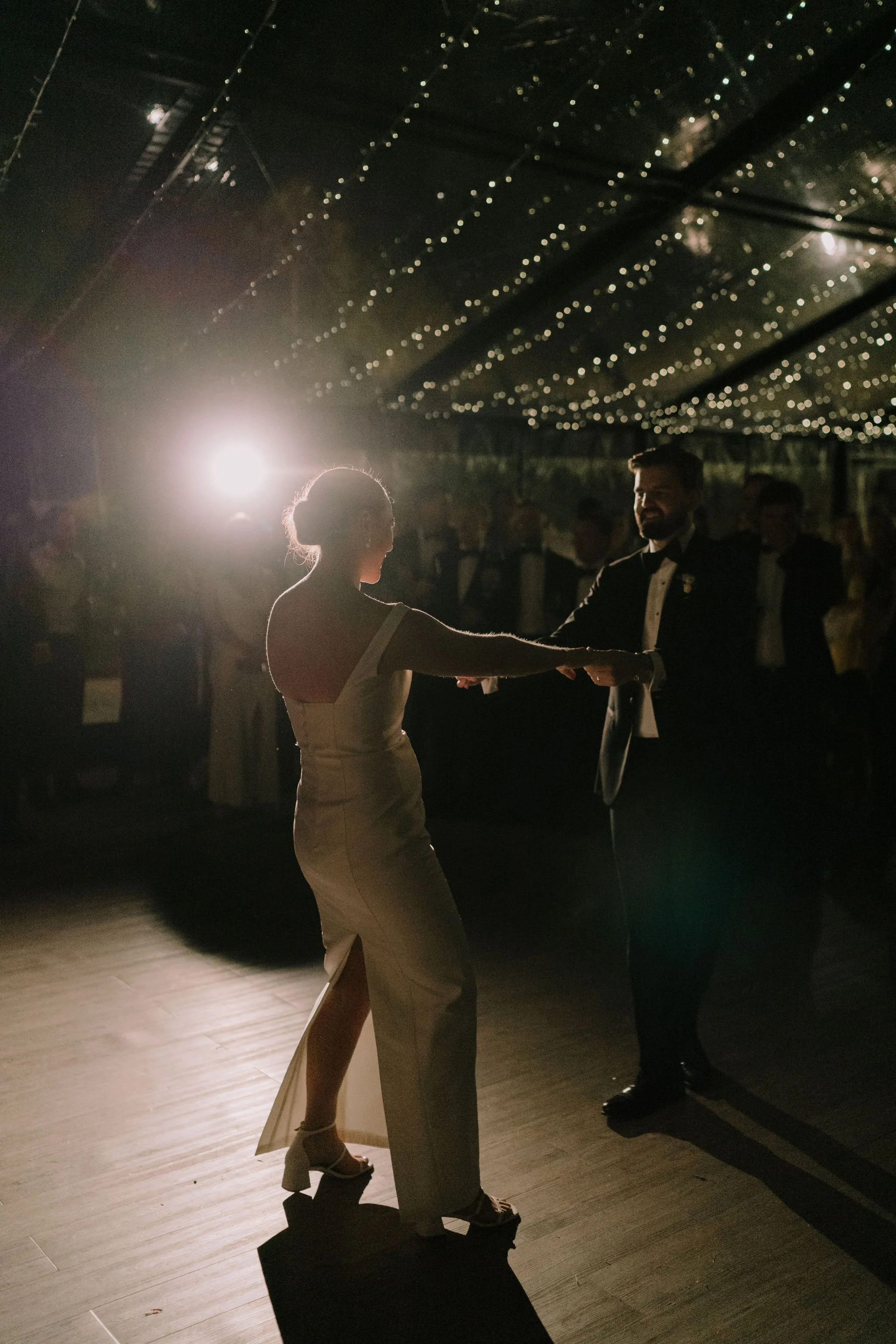 A bride and groom dancing during their wedding reception, with the bride in a white gown and the groom in a tuxedo, under a ceiling decorated with string lights, as guests watch in the background.
