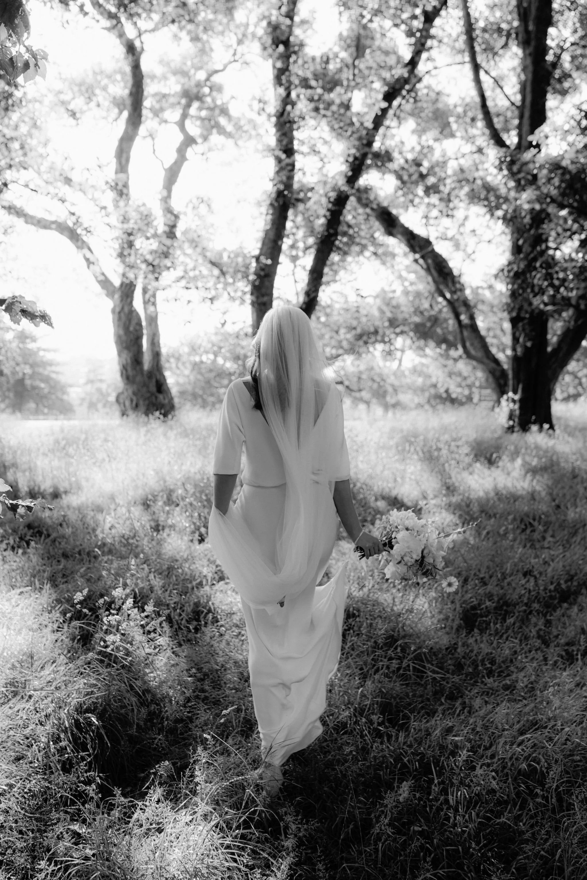 A woman in a flowing white dress walking through a sunlit forest holding a bouquet of flowers.