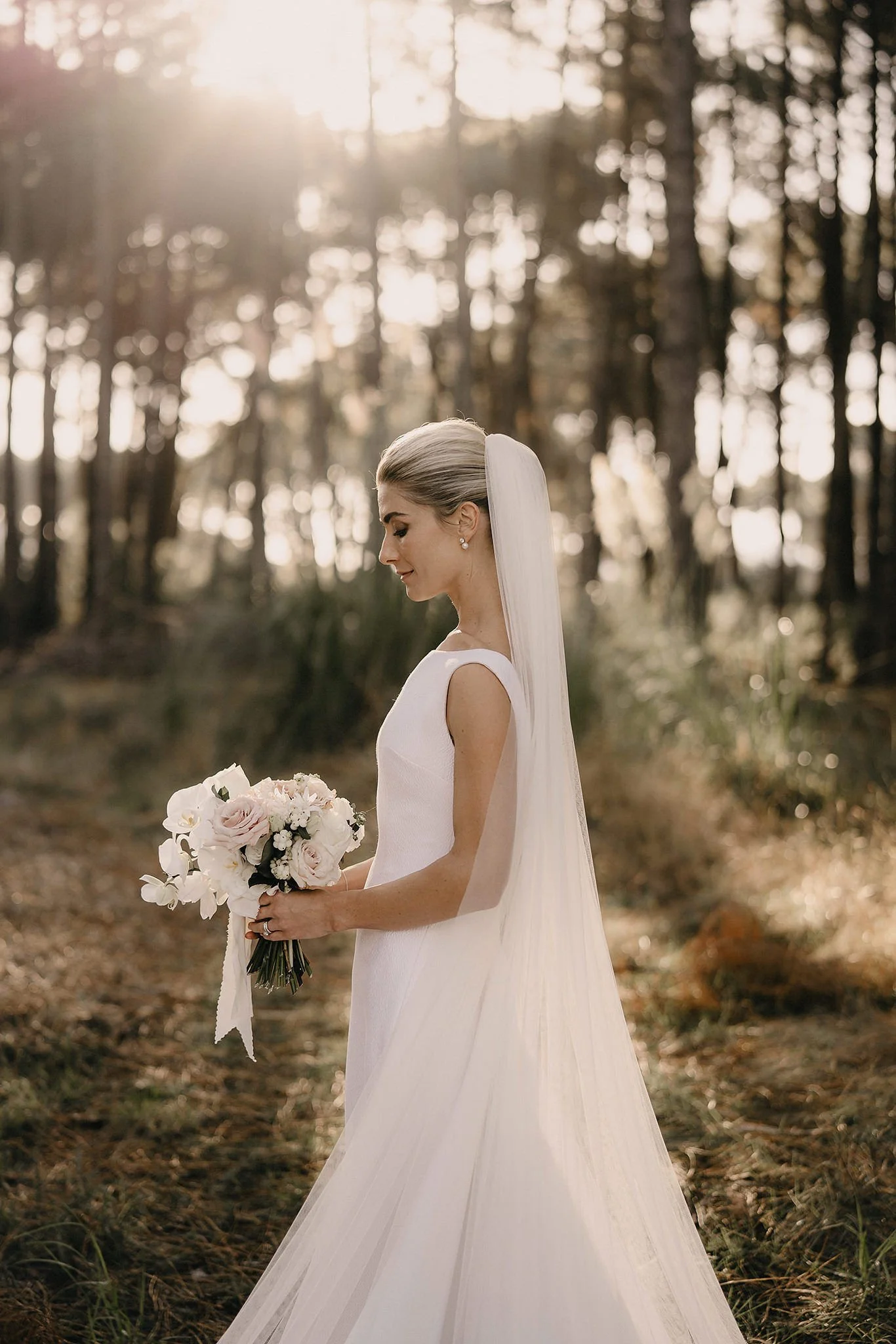 A bride with blonde hair styled in a sleek updo and wearing pearl earrings is standing outdoors in a wooded area during sunset, holding a bouquet of white and blush pink flowers.