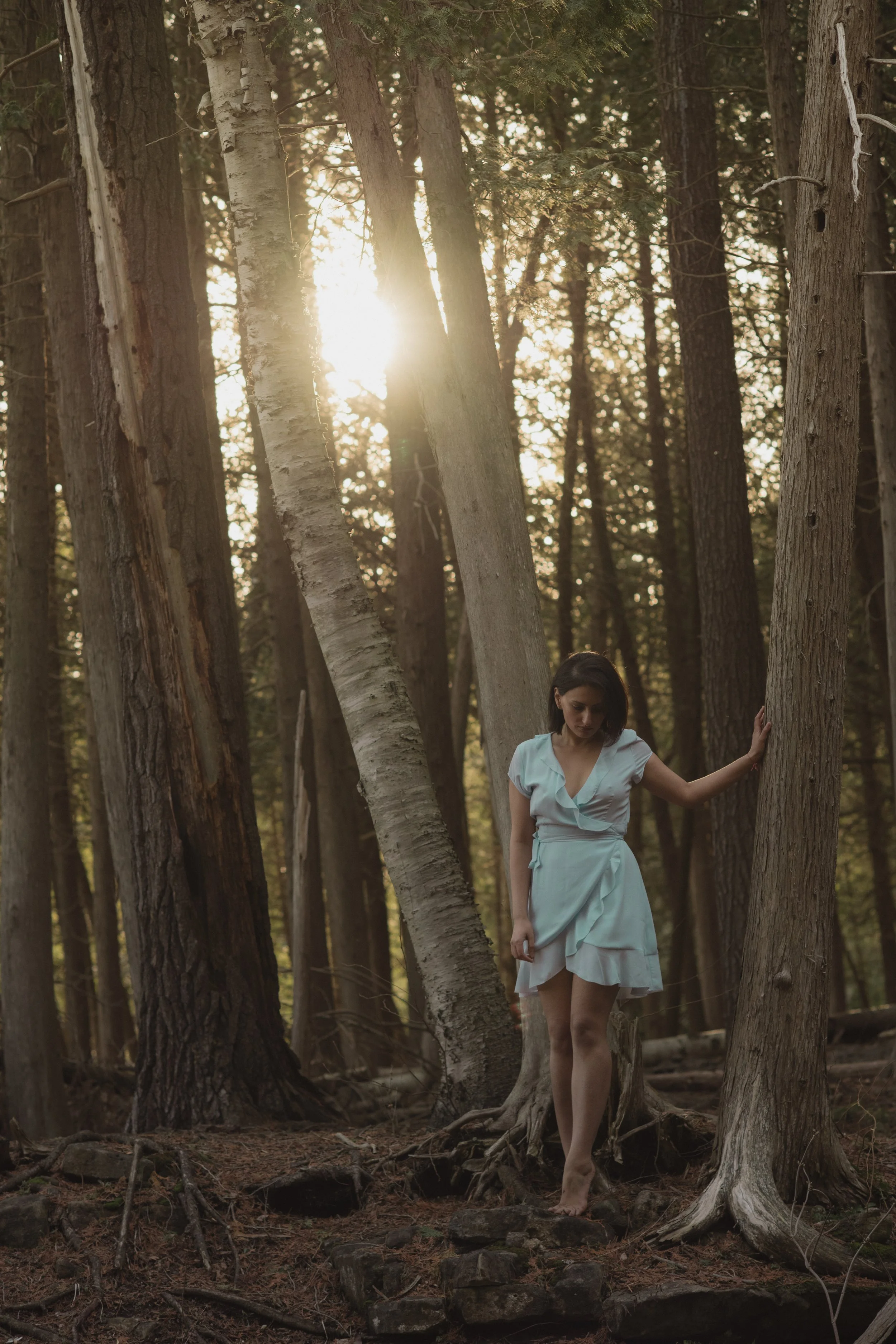 A woman in a pale blue dress standing barefoot in a forest, touching a tree, with sunlight filtering through the trees.