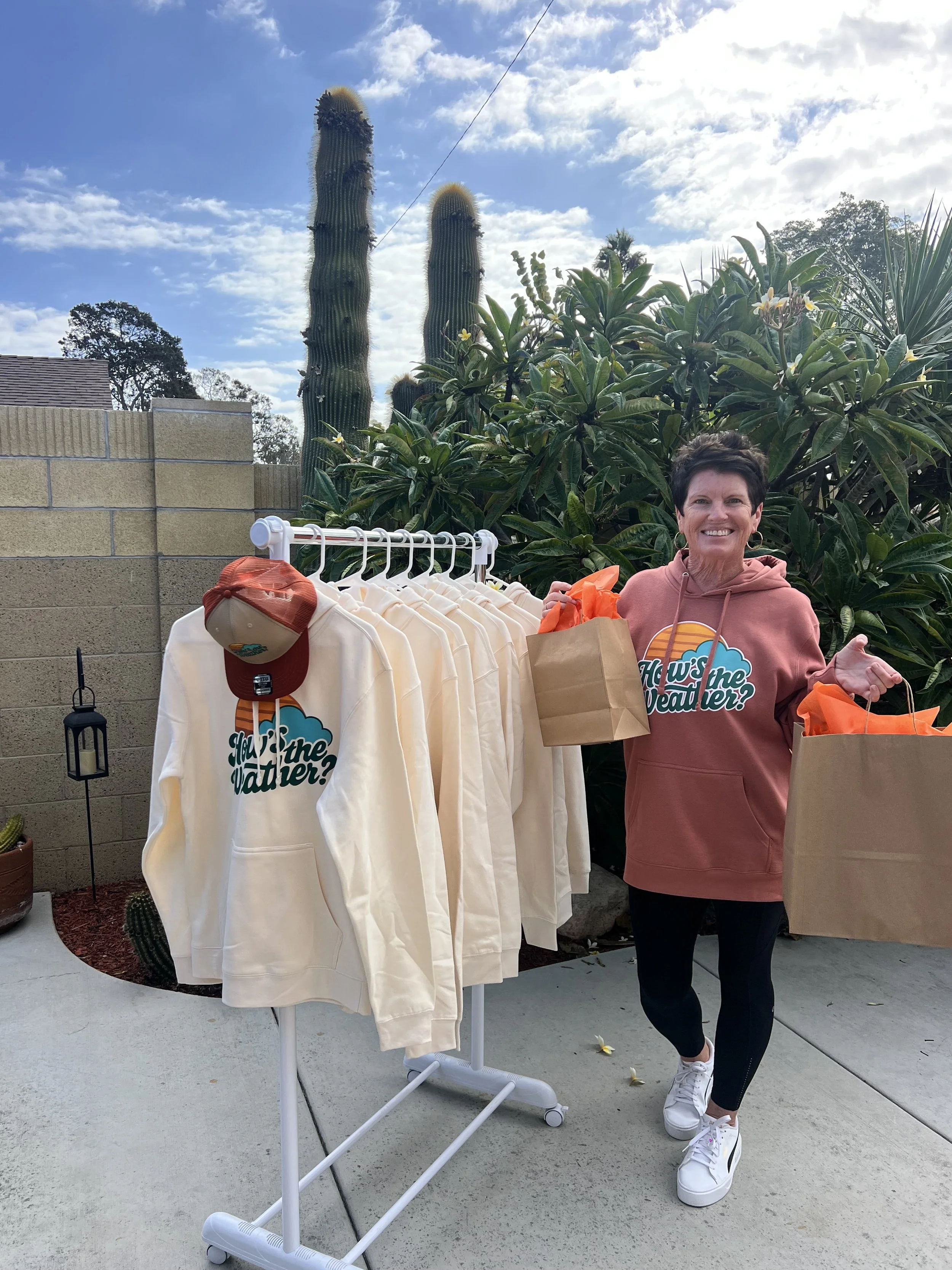 A photo of Erin Clark standing next to a rack of her sweatshirts that say, "How's the Weather?" 