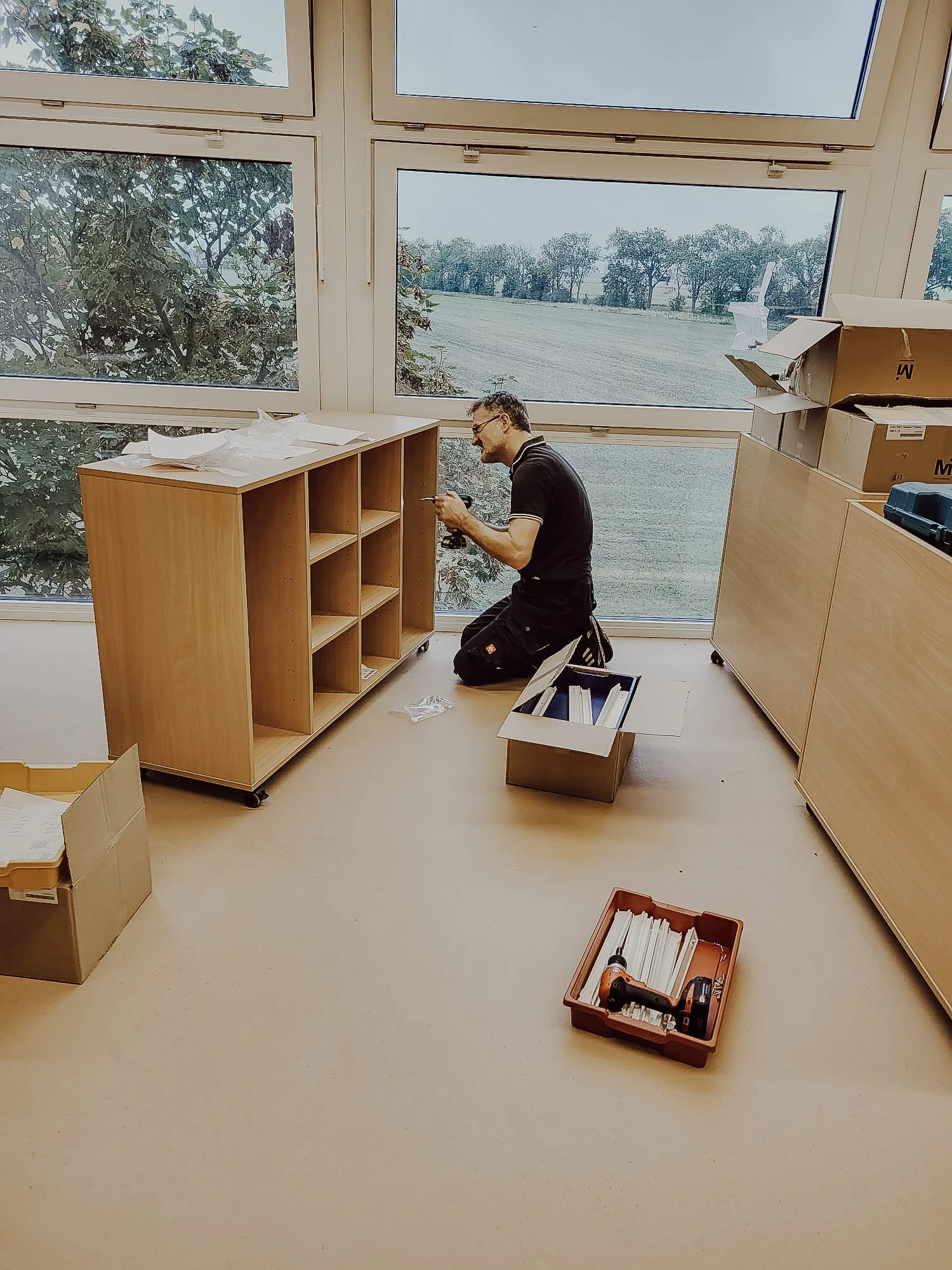 Ein Mann arbeitet in einem hellen Raum mit großen Fenstern, die eine ländliche Landschaft zeigen. Er sitzt auf einem Hocker neben einem Holzregal, das noch zusammengebaut wird, mit Werkzeugen und Verpackungsmaterialien um ihn herum.