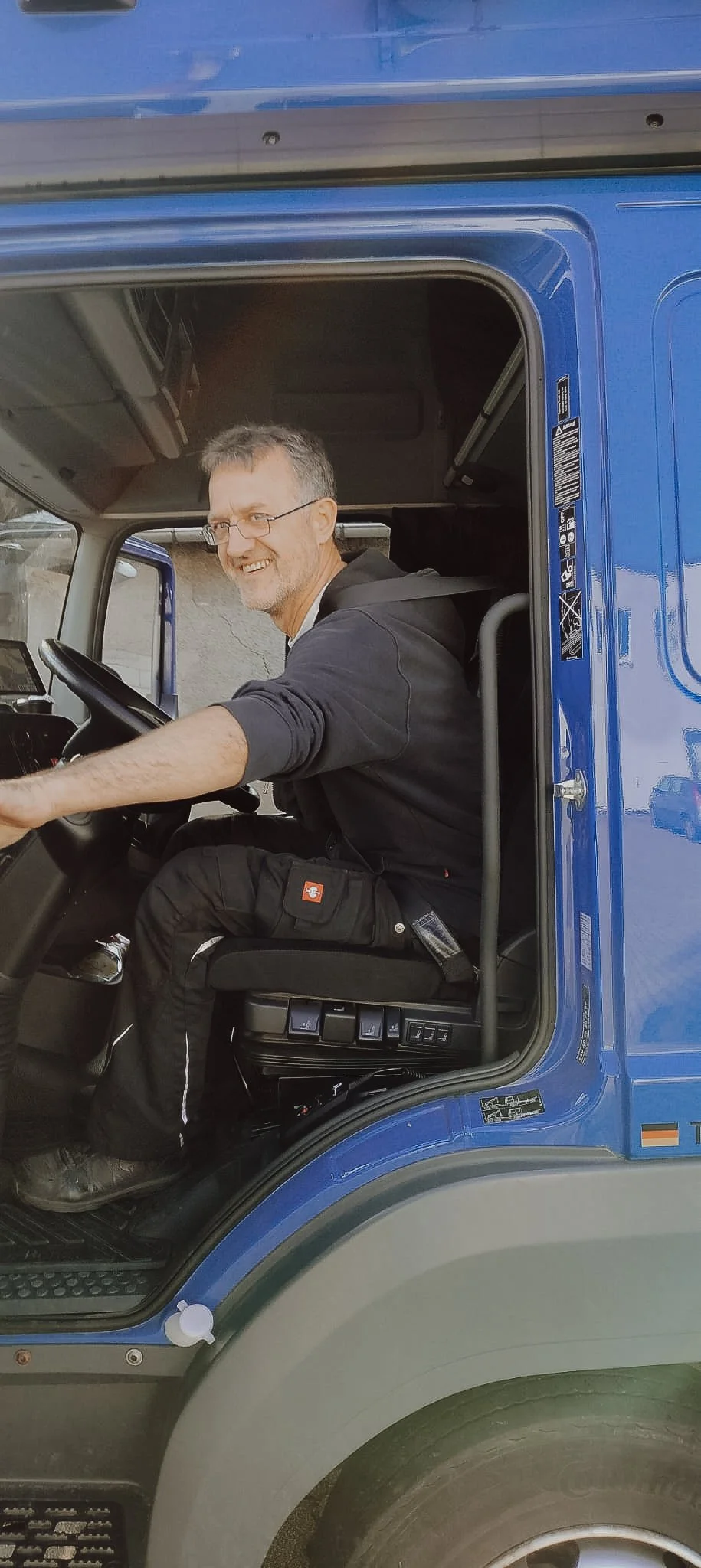 Ein Mann sitzt in der Fahrerkabine eines blauen Lastwagens, lächelt und schaut zur Kamera.