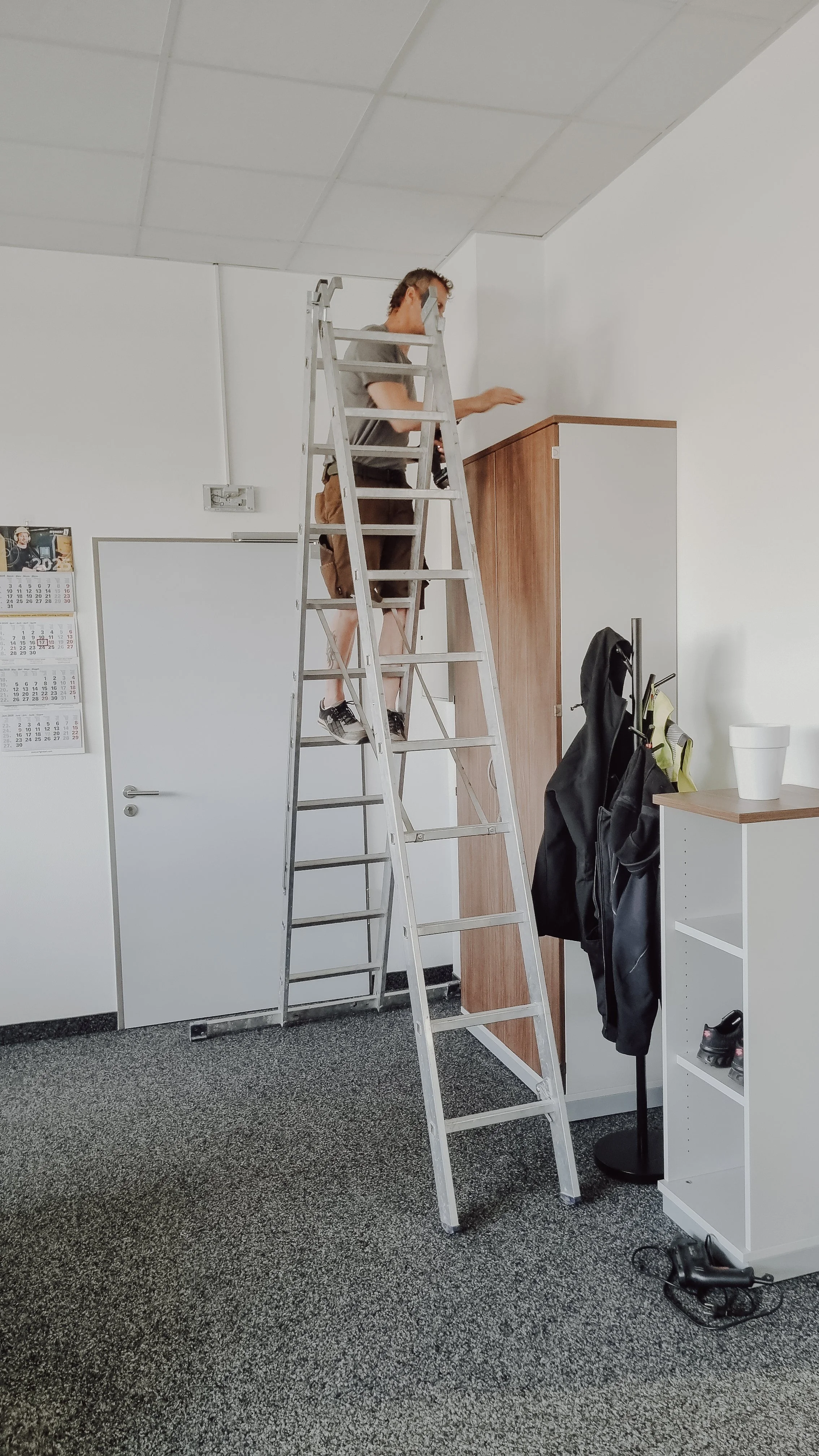 Ein Mann steht auf einer Leiter und arbeitet an der Decke eines Raumes, während er einen weißen Raum mit weißen Wänden und einer grauen Teppichboden hat.