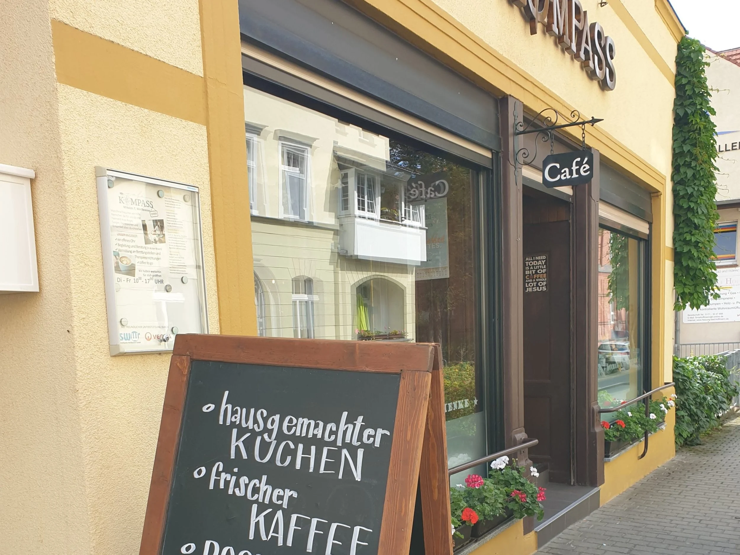 Außenansicht eines Cafés mit schwarzem Hinweisschild, das auf der Tafel vor der Tür 'hausgemachter Kuchen, Kaffee, frischer Kaffee' schreibt. Das Café befindet sich in einem gelben Gebäude mit großen Fenstern und bunten Blumen vor dem Eingang.