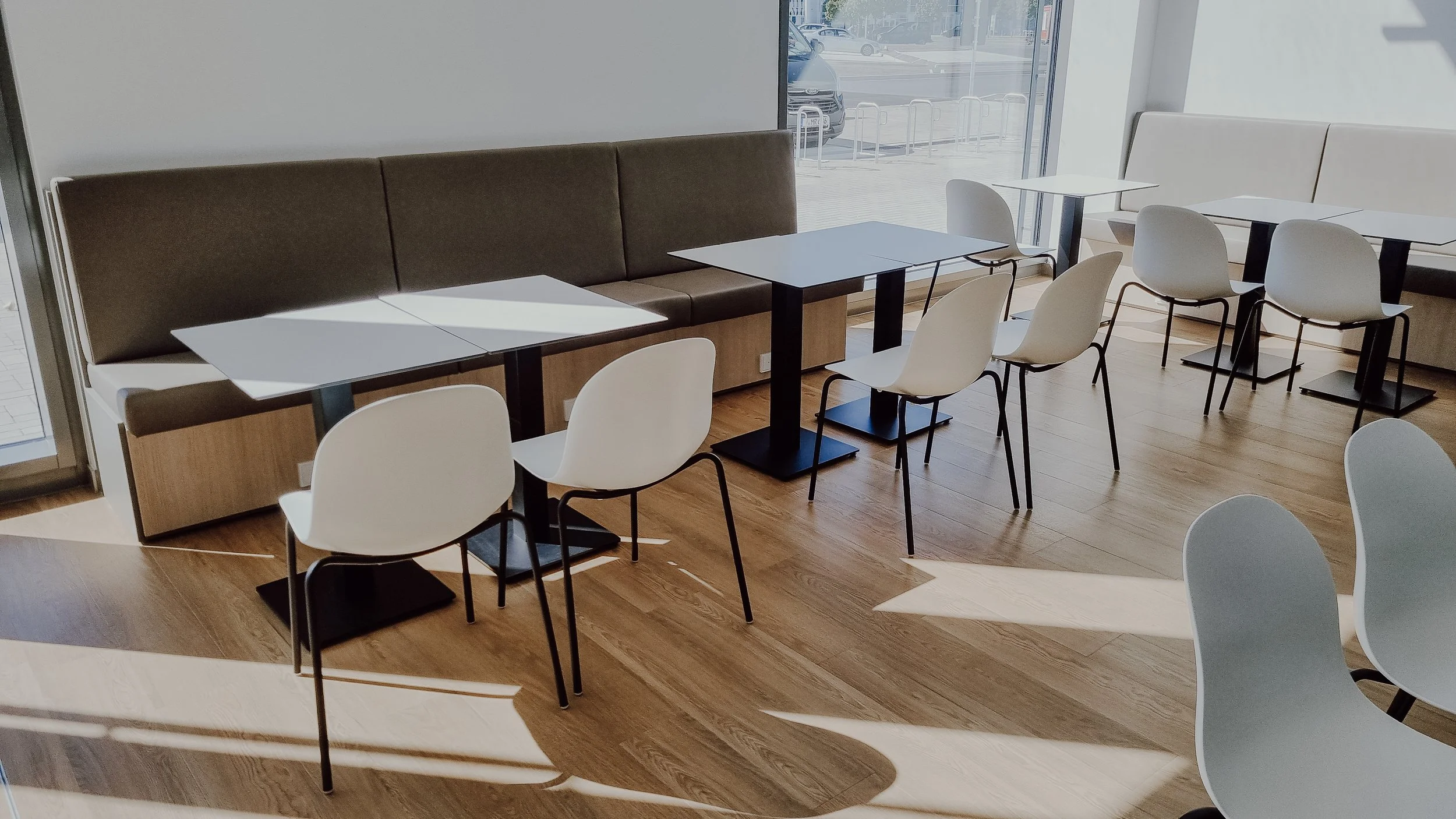 Leere Sitzgelegenheiten in einem modernen Café mit großen Fenstern, Holzboden und weißen Tischen und Stühlen.
