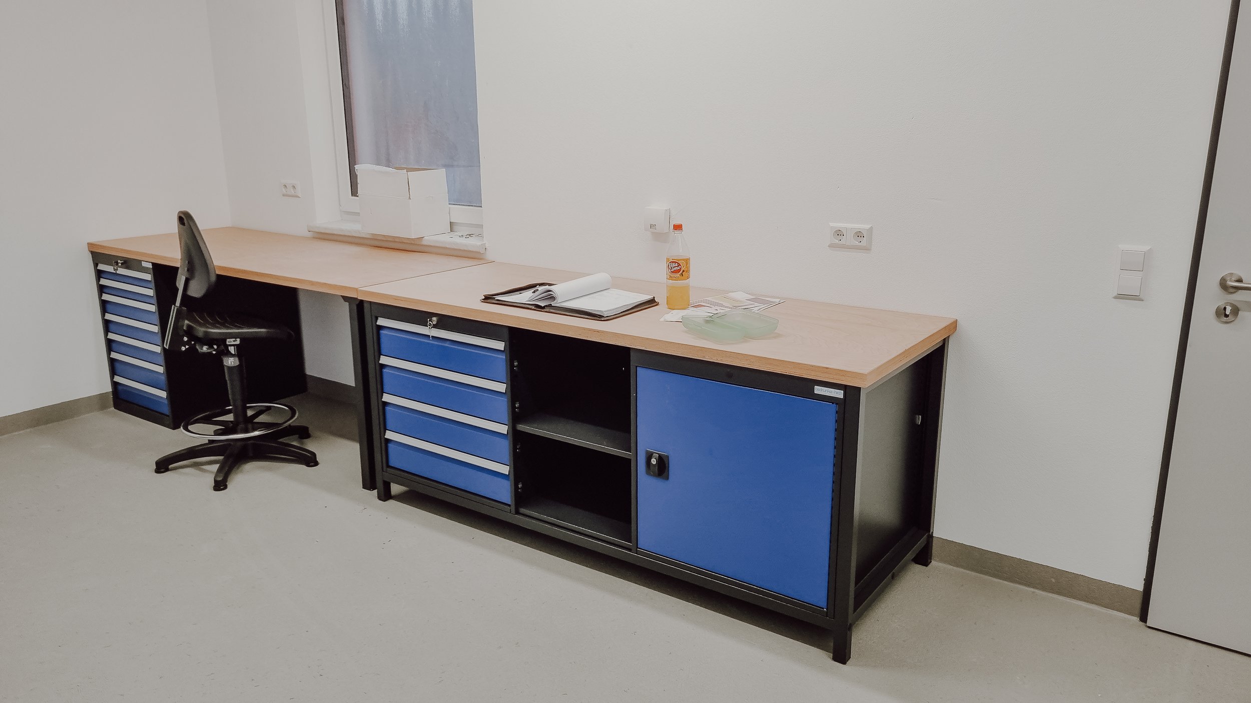 Ein leeres Büro mit einem schwarzen Schreibtisch, blauen Schubladen, einem schwarzen Bürostuhl, einem Fenster, einem geöffneten Buch, einer Flasche Desinfektionsmittel, Papier und andere Gegenstände auf dem Tisch.