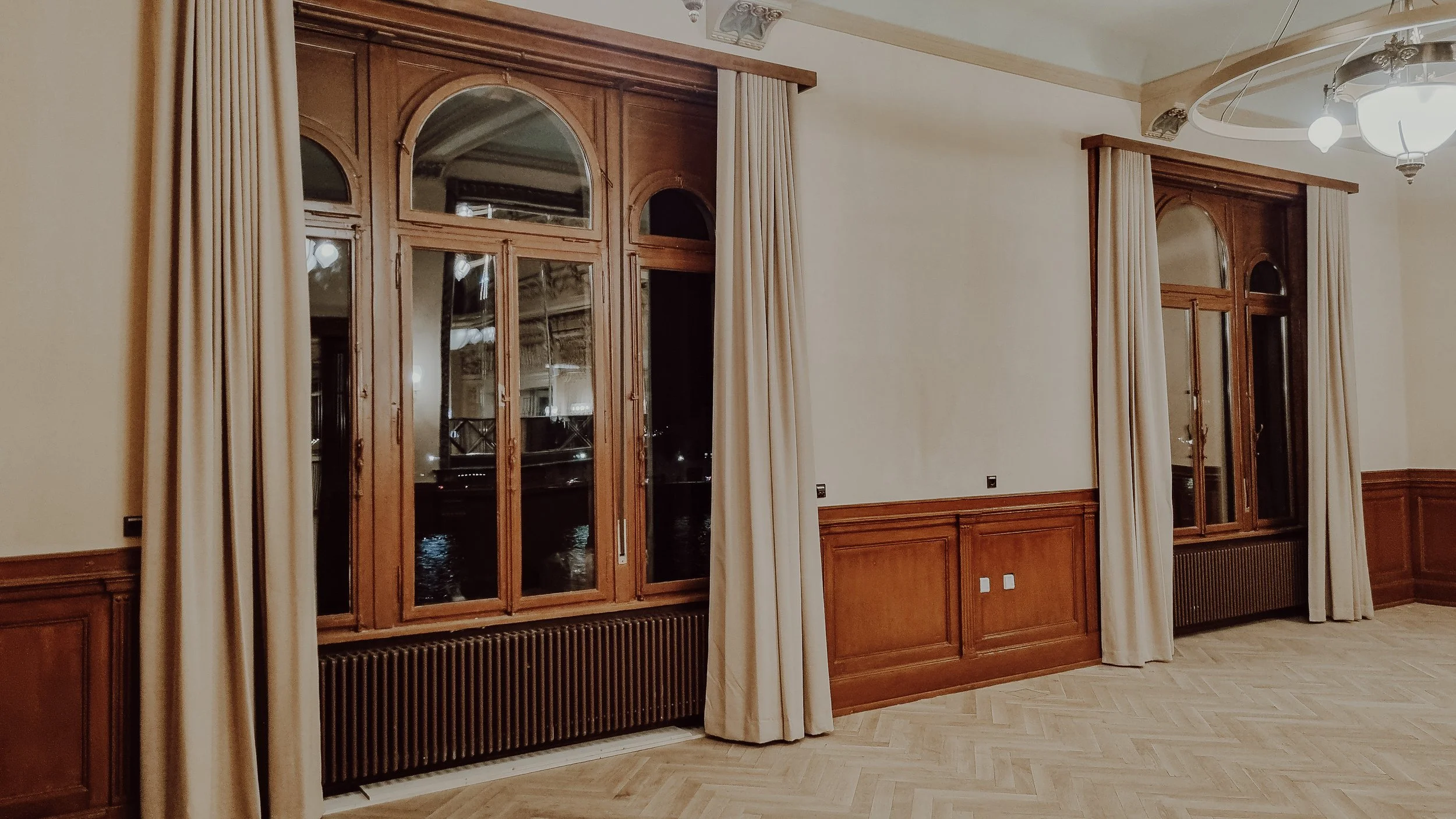 Ein Raum mit großen Fenster, Holzrahmen, und weißen Vorhängen, an der Wand. Es gibt eine Deckenleuchte und Holzwände, Parkettboden.
