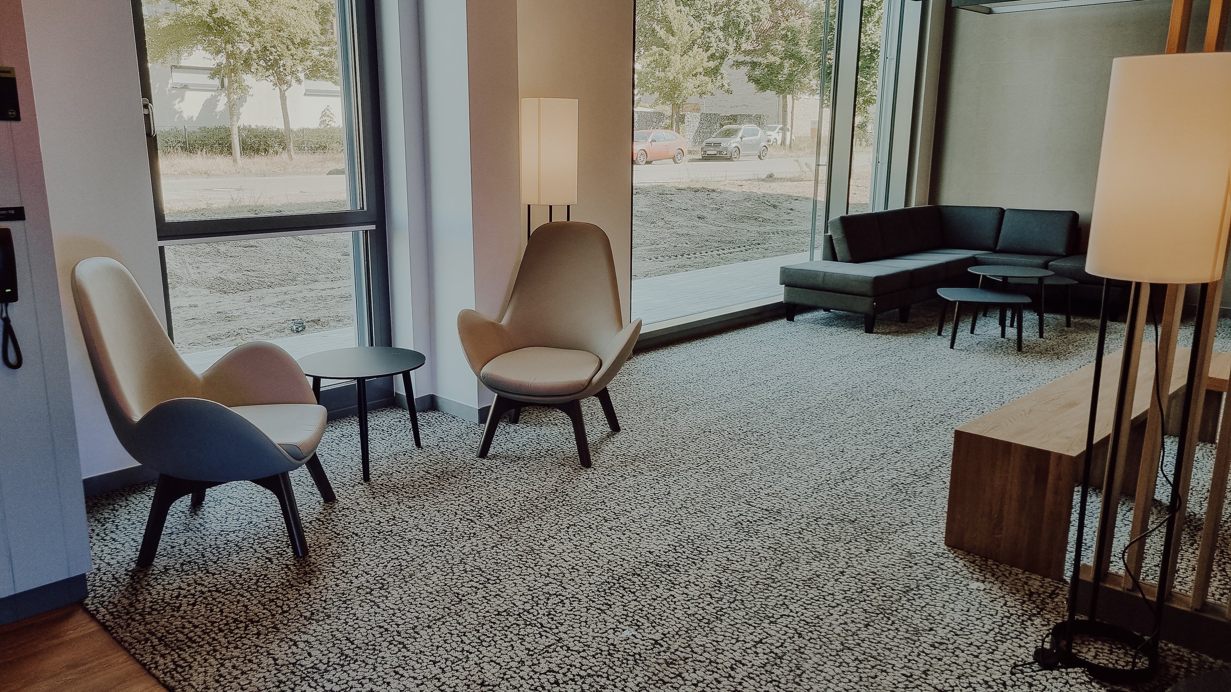 Ein modern eingerichteter Loungebereich mit Sesseln, einem Sofa, Beistelltischen, Stehlampen und großen Fenstern, durch die man auf eine Straße mit geparkten Autos und Bäumen blickt.