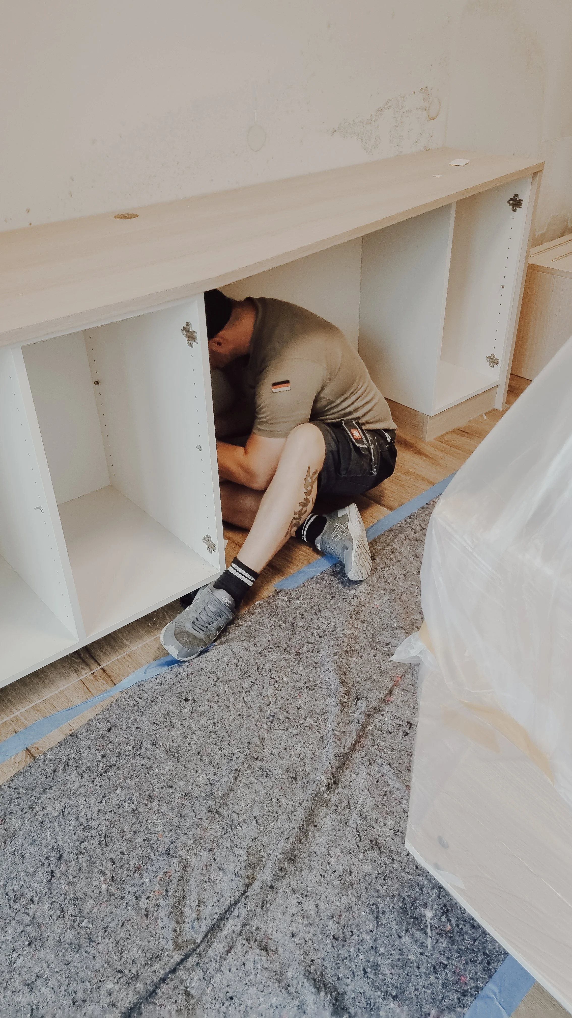 Mann baut eine weiße Holz-Schrankkombination in einem Raum, sitzt auf dem Boden, trägt ein graues T-Shirt und Shorts, arbeitet an einem der Schränke, während eine Plastikplane und eine Teppichrolle im Raum liegen.