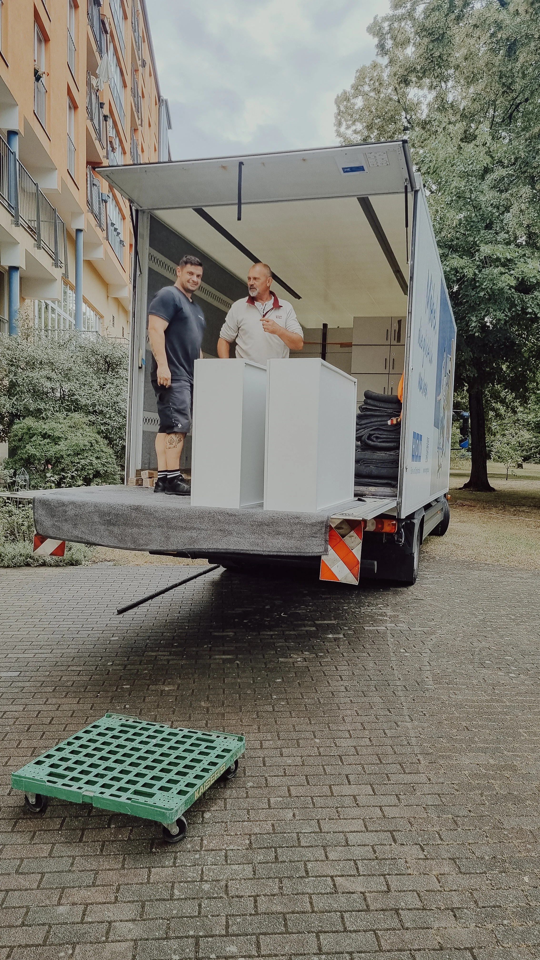 Zwei Männer stehen in einem offenen Lieferwagen, der vor einer Mehrfamilienwohnanlage geparkt ist. Der Lieferwagen ist voll mit Möbeln, darunter ein weißer Tisch und ein Paketsack. Eine grüne, rollbare Kunststoffpalette liegt auf dem Boden vor dem Wa