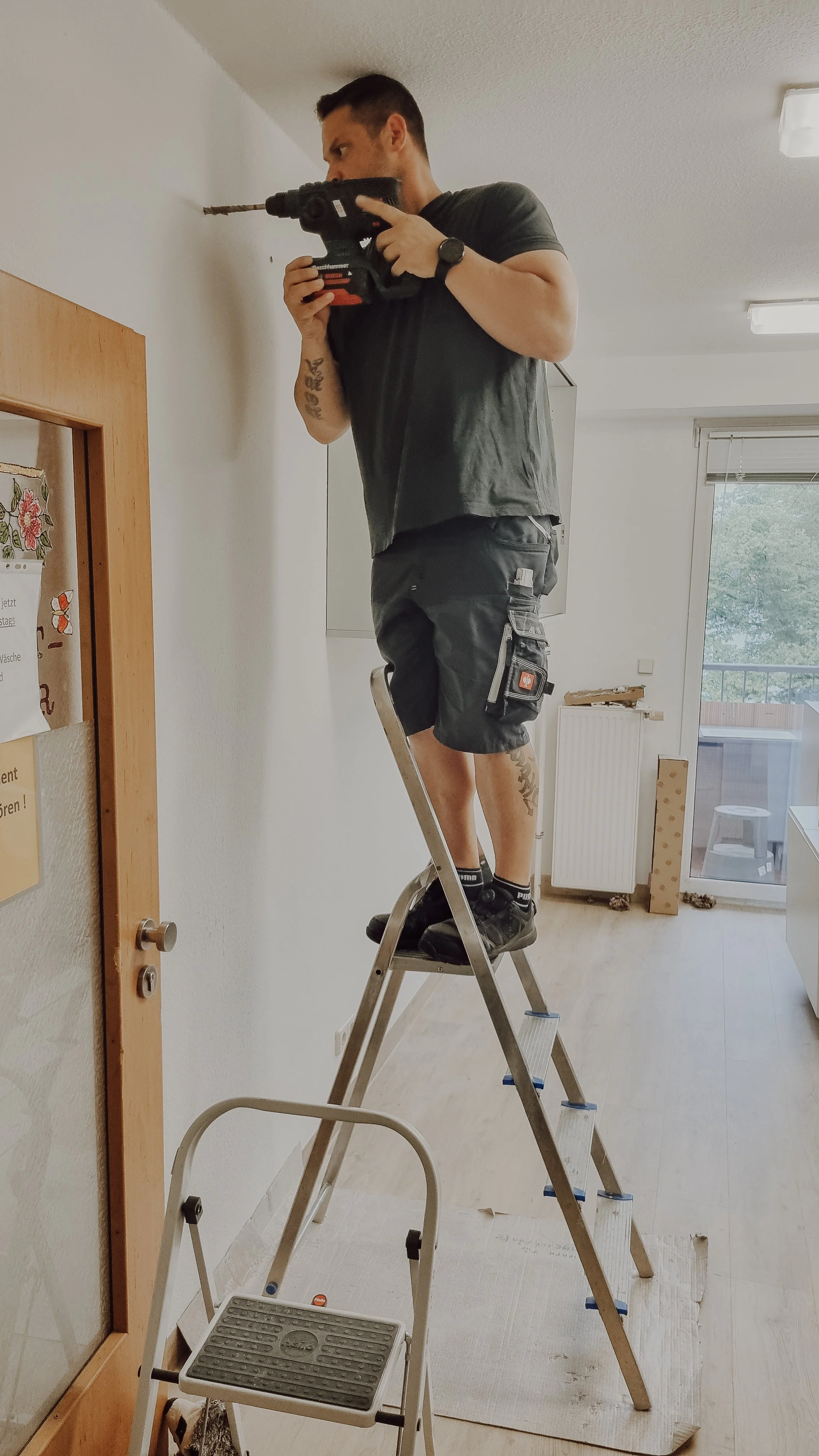 Ein Mann steht auf einer Leiter, um eine Wand zu reparieren oder zu streichen, während er ein Bohrgerät benutzt.