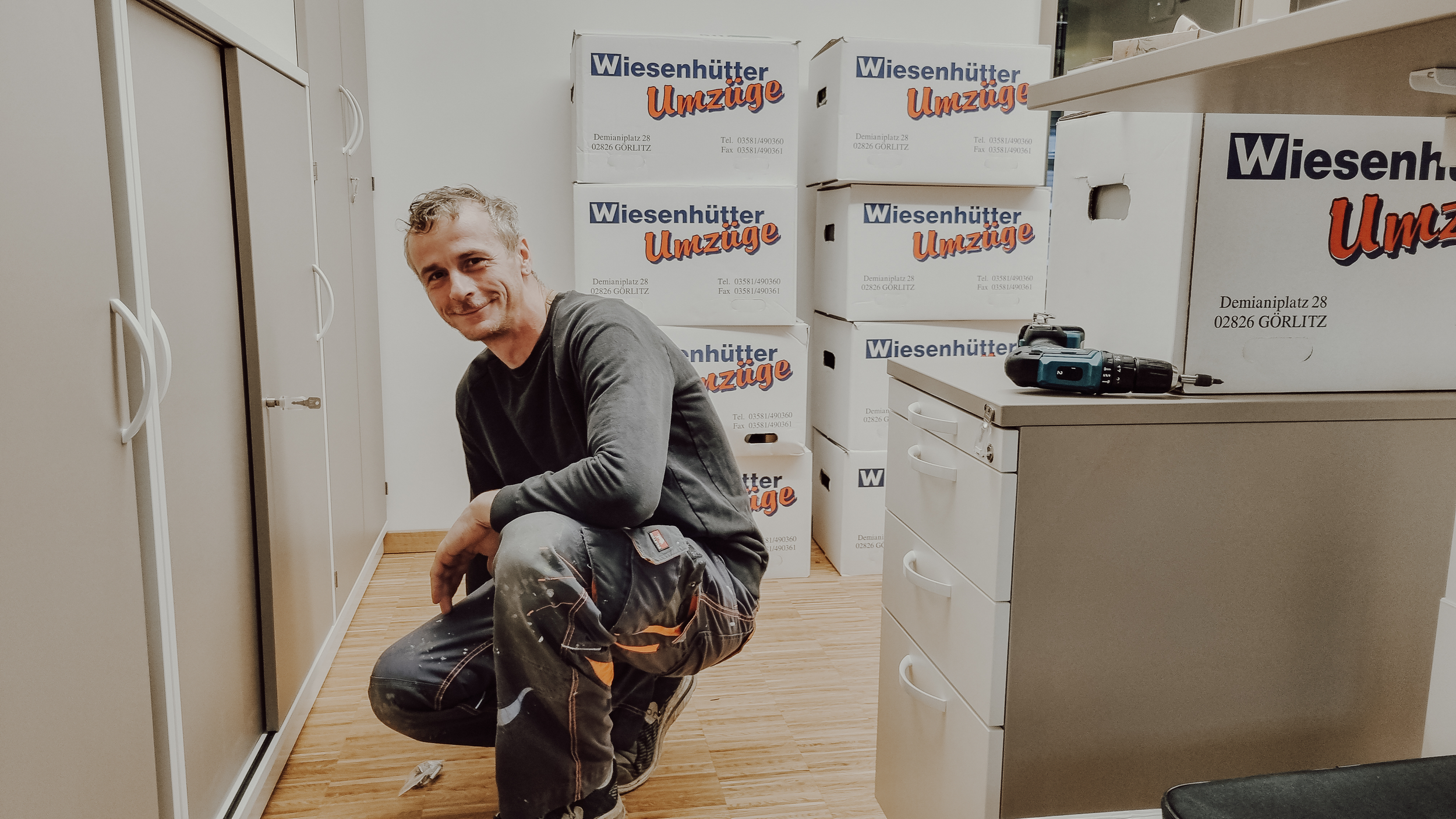 Ein Mann ist in einem Raum mit Umzugskartons, die mit 'Wiesenhütter Umzüge' beschriftet sind. Er trägt Arbeitskleidung und kneift in eine Wand. Daneben befinden sich Werkzeuge und weiße Schränke.