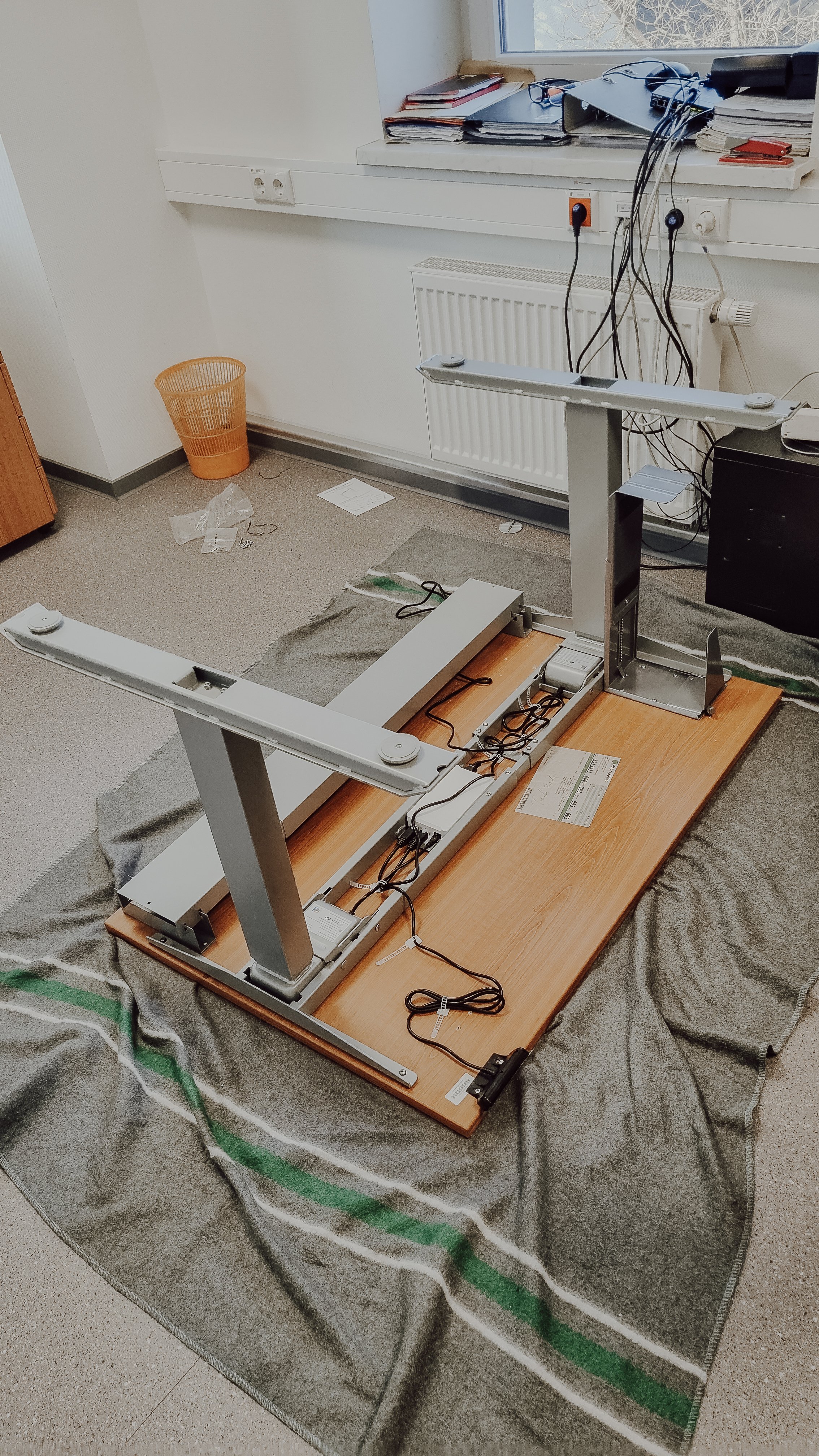 Ein auseinandergebauter Tisch auf einem Teppichboden in einem Büro, mit Kabeln und Elektronikkomponenten. Im Hintergrund eine Fensterbank mit Papieren und Büromaterialien.