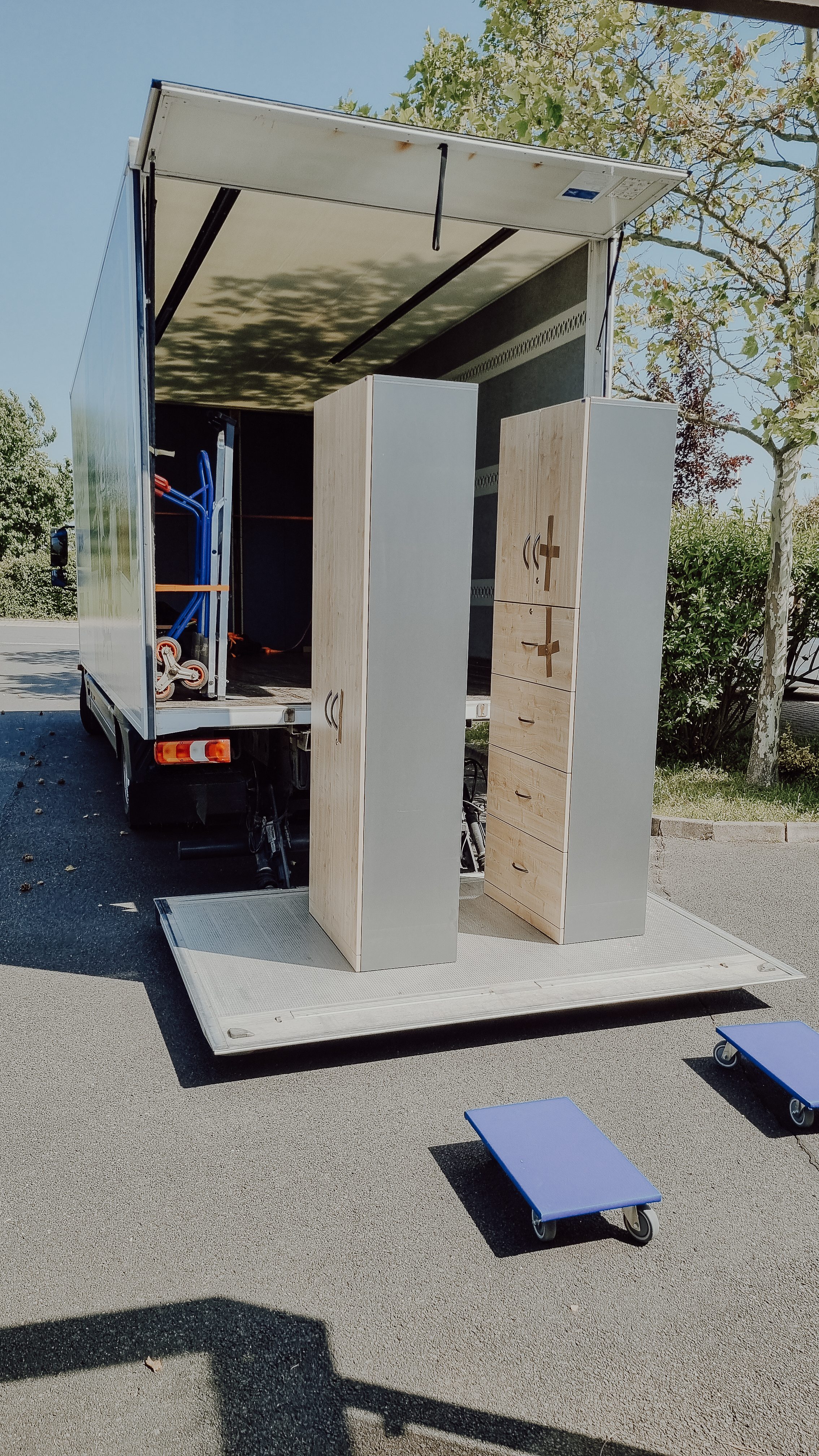 Ein beladener Umzugskarton mit zwei zurechtgestellten Schränken und kleinen Rollbrettern auf Asphalt im Freien. Im Hintergrund sind Bäume und ein blauer Himmel sichtbar.