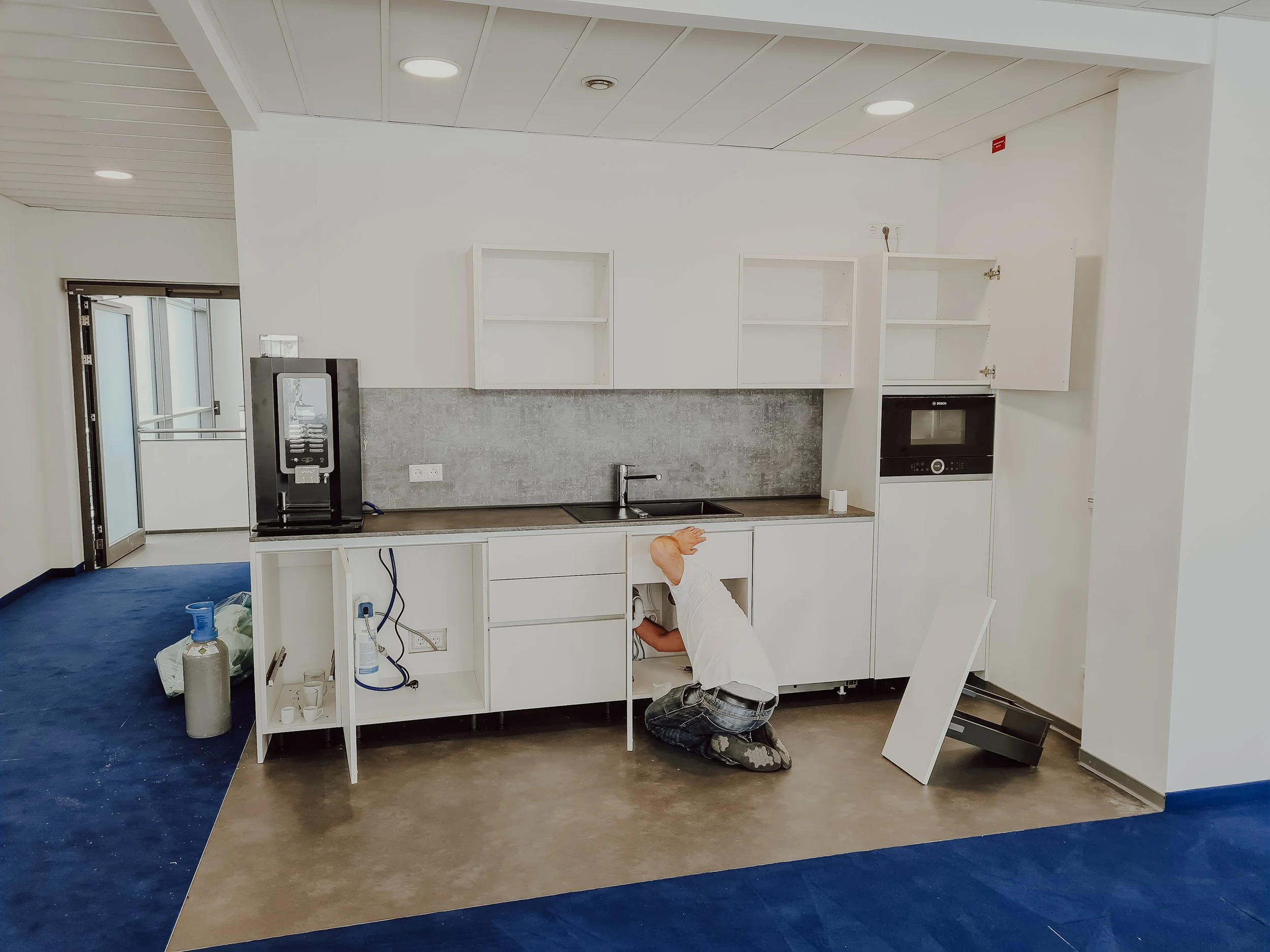 Eine Person arbeitet an einem Küchenumbau und installiert weiße Küchenschränke. Die Arbeitsfläche ist teilweise fertig, und es befinden sich Bauteile sowie Werkzeuge und Materialien um die Arbeitsstelle herum.