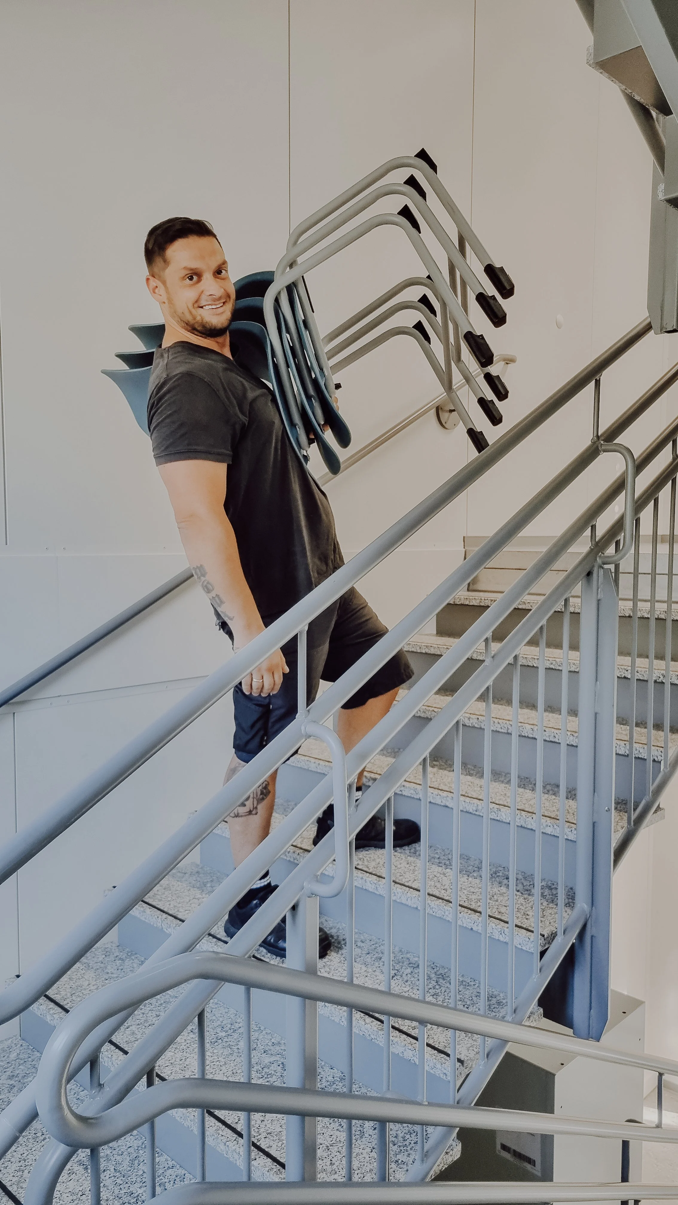 Ein Mann trägt mehrere Stühle an einem Handlauf die Treppe hinauf und lächelt in die Kamera.