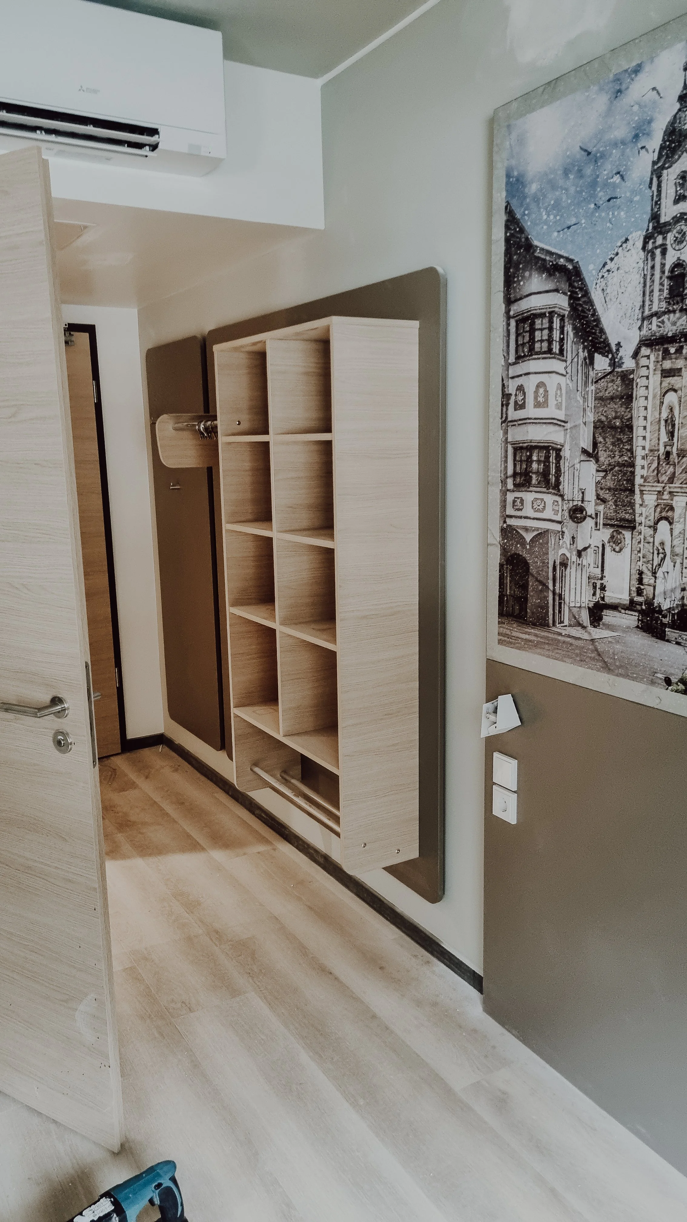 Ein Raum mit einer weißen Wand, auf der ein Wandgerät installiert ist, und einem offenen, leeren hölzernen Regalschrank. Es gibt eine helle Holztür auf der linken Seite und eine Fotografie einer Stadtansicht an der Wand.