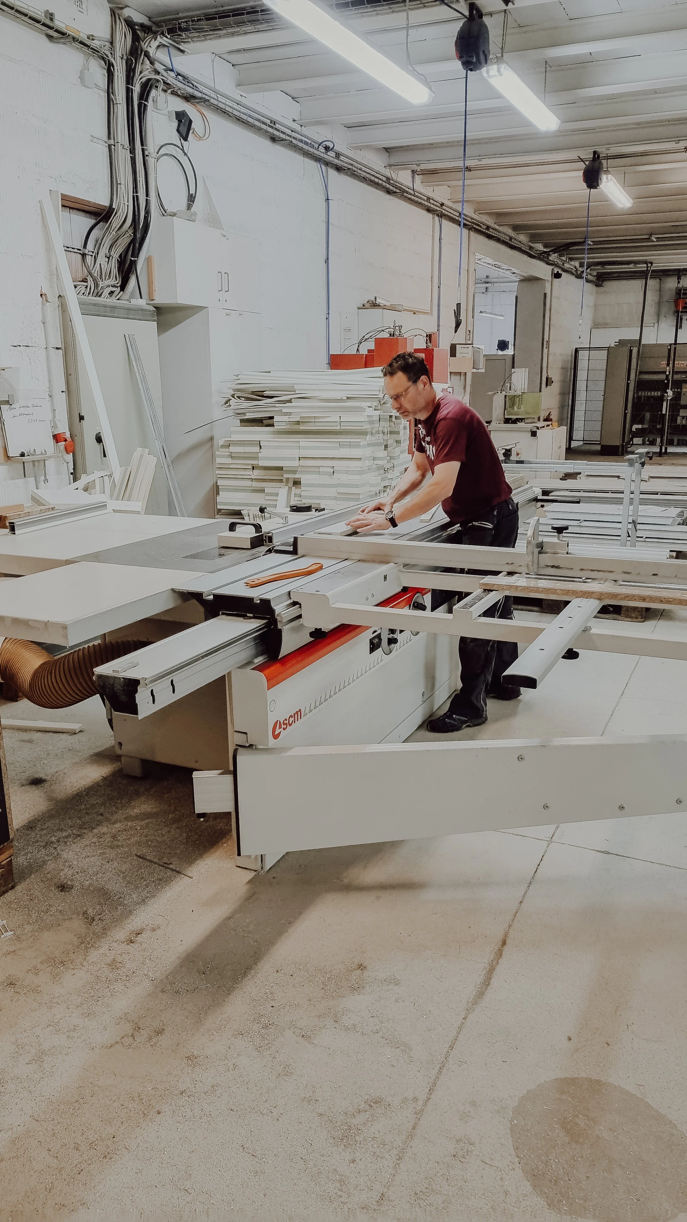 Mann arbeitet an einer Sägemaschine in einer Werkstatt mit Holzplatten im Hintergrund.