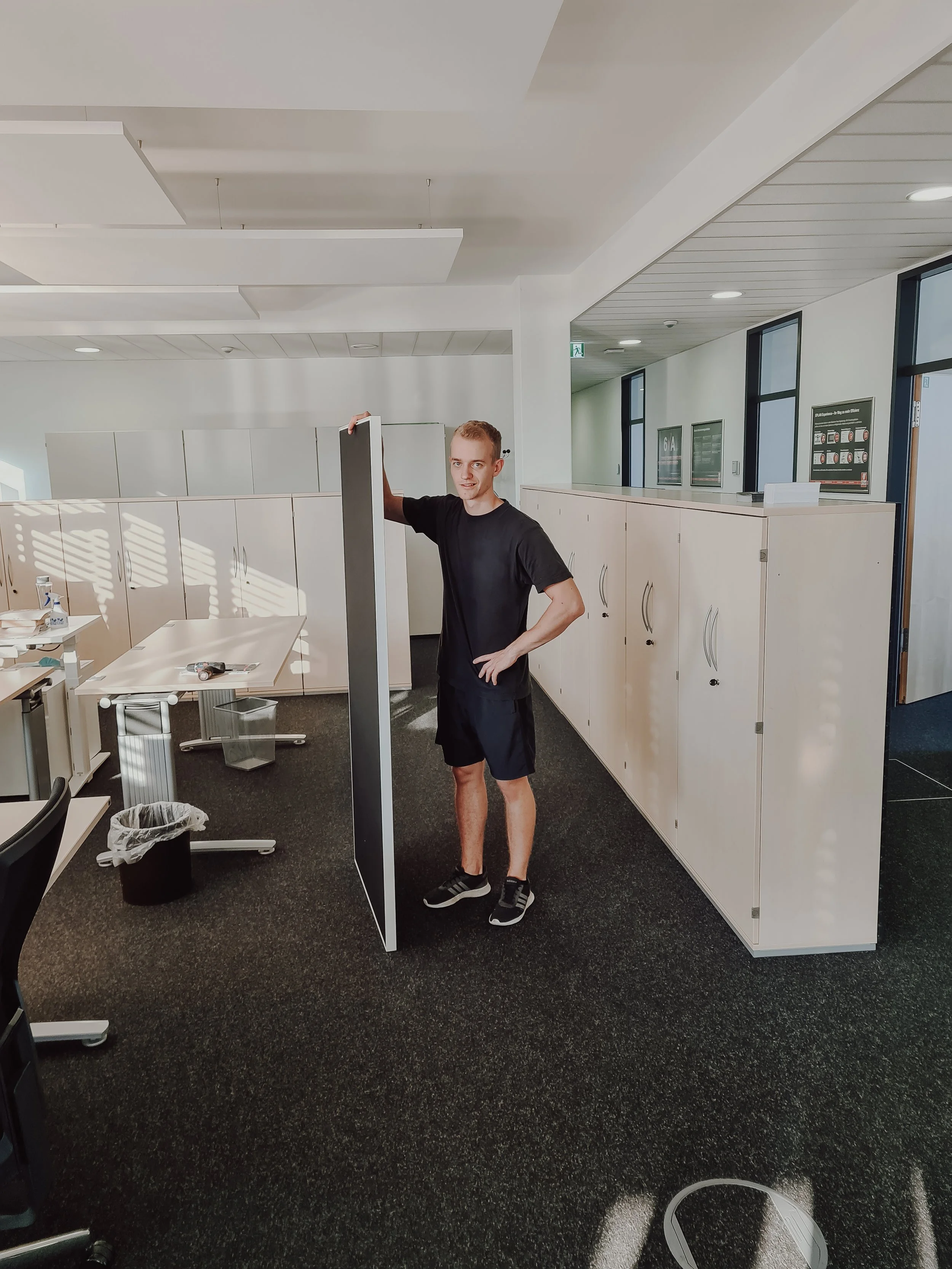 Junge Mann in schwarzem T-Shirt und Shorts steht in einem Büroraum neben einem Trennwand, umgeben von weißen Schränken, einem Tisch, und einem Papierkorb. Der Raum ist gut beleuchtet mit Fenstern und Deckenleuchten.