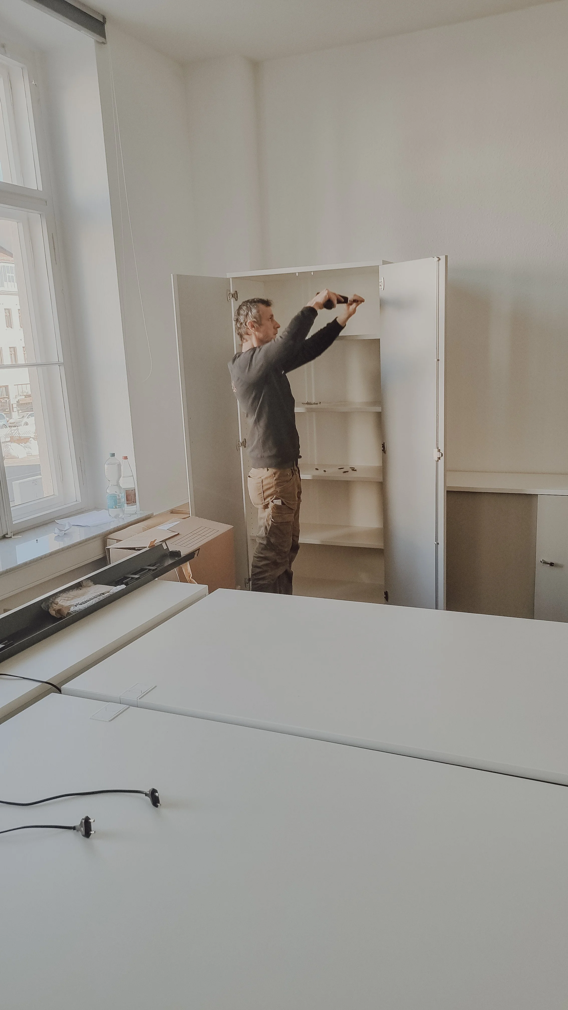 Ein Mann repariert eine weiße Küchenschranktür in einem hellen Raum mit einem Fenster an der Seite.