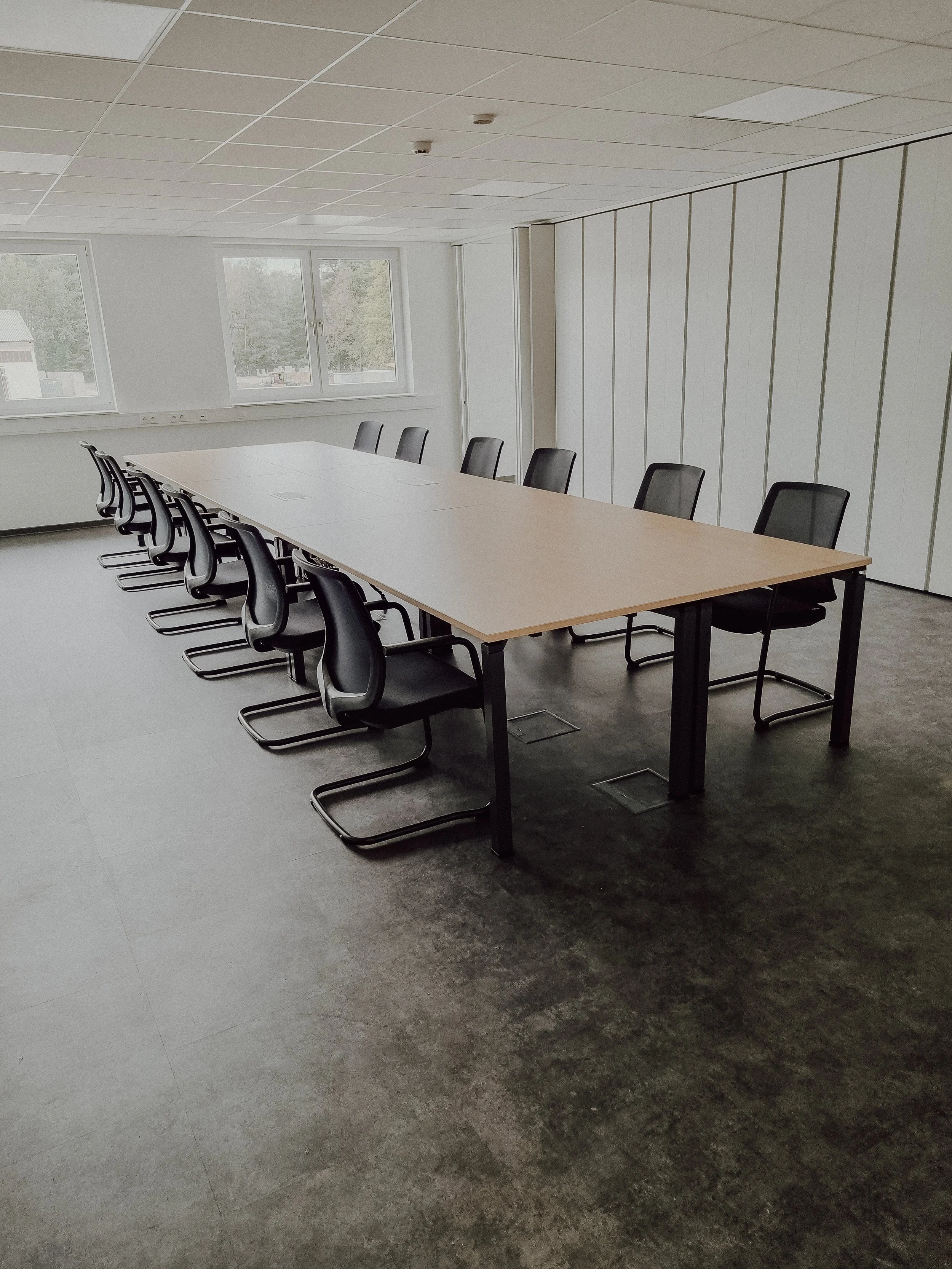 Ein leeres Konferenzraum mit einem langen Tisch und mehreren schwarzen Stühlen. Es gibt Fenster, die Blick auf die Außenwelt bieten, und die Wände sind weiß. Der Raum hat eine schlichte, professionelle Atmosphäre.