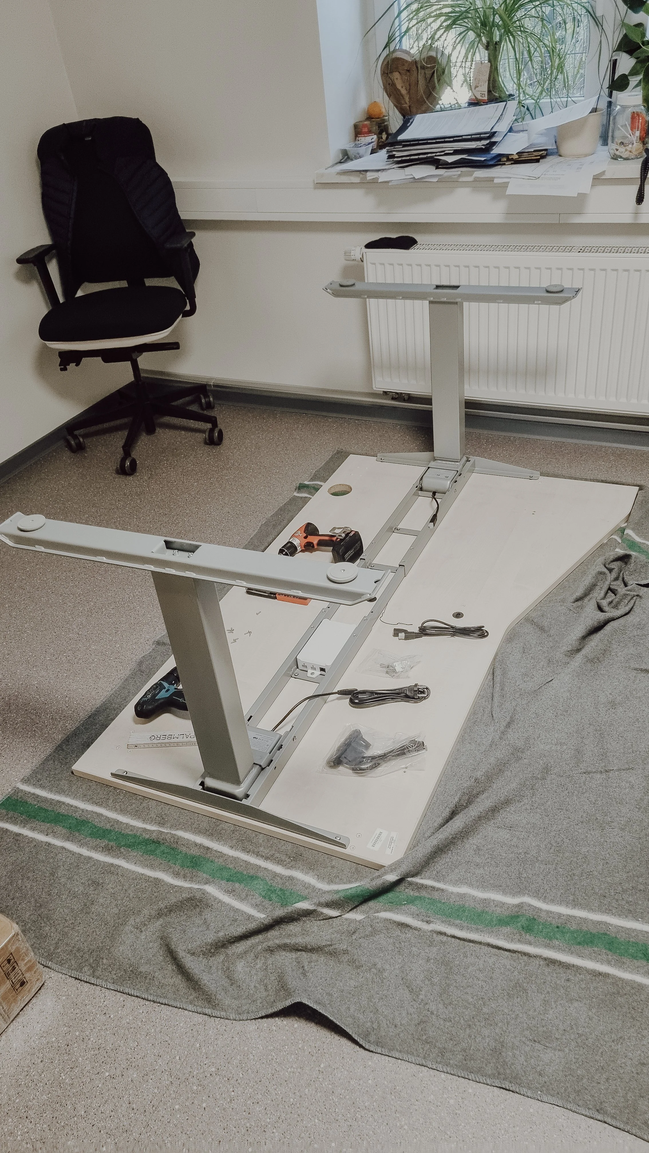 Ein Schreibtisch mit einem schwarzen Bürostuhl, auf dem eine schwarze Jacke liegt, und einem Tisch, der teilweise aufgebaut wird. Im Hintergrund befindet sich eine Fensterbank mit Pflanzen und Papieren.