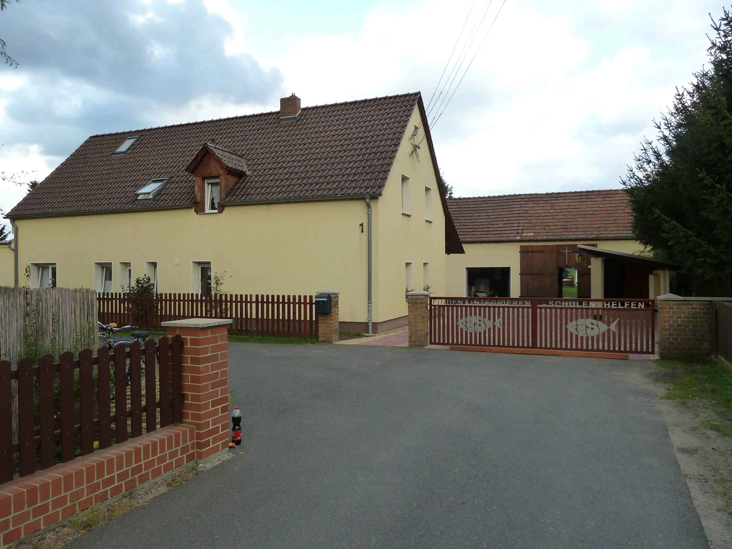 Ein gelbes Haus mit teilweise roten Ziegeldächern und mehreren Dachfenstern, umgeben von einem Zaun und Bäumen, bei bewölktem Himmel.