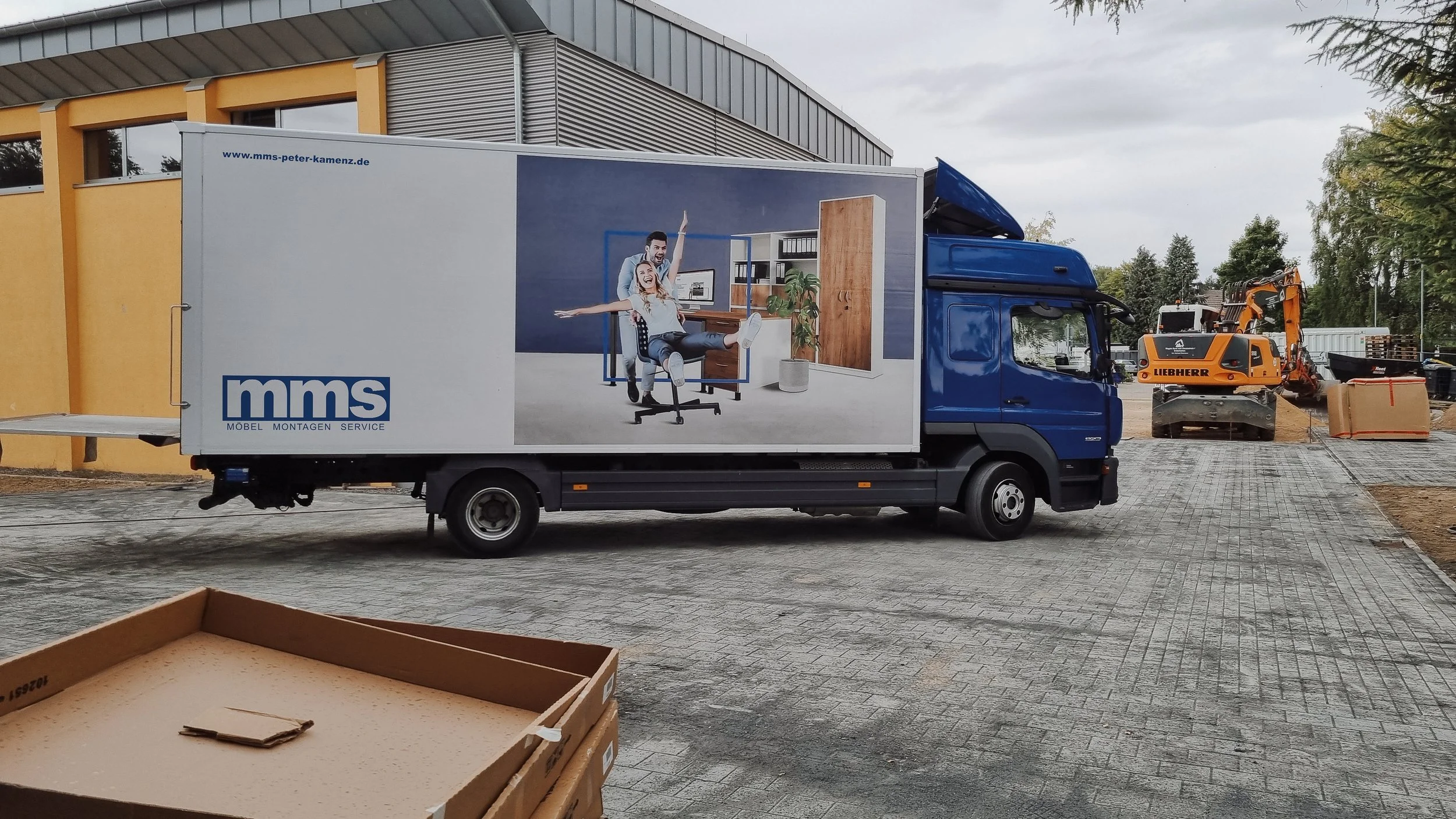 Ein umgebauter LKW mit Werbung für Möbeldienstleistungen, auf einer Baustelle mit Bagger und Baugeräten im Hintergrund.