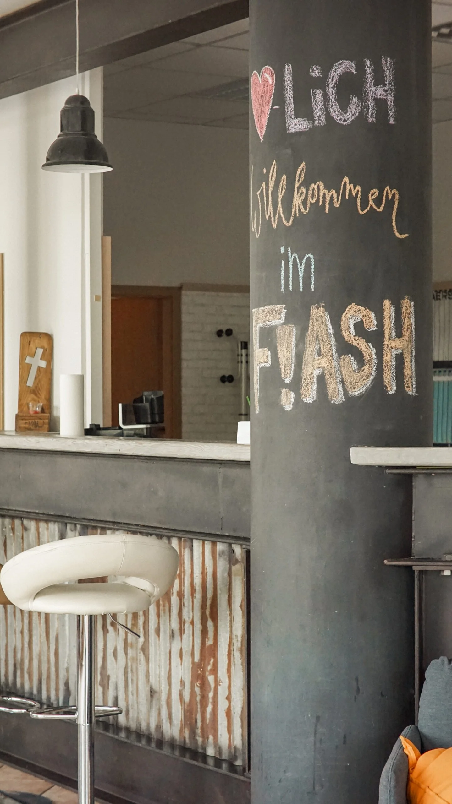 Ein schwarzes Tafel mit bunten Kreidebuchstaben, die 'LICH', 'Willkommen', 'im' und 'F!ASH' lesen, in einem Café oder Bistro.