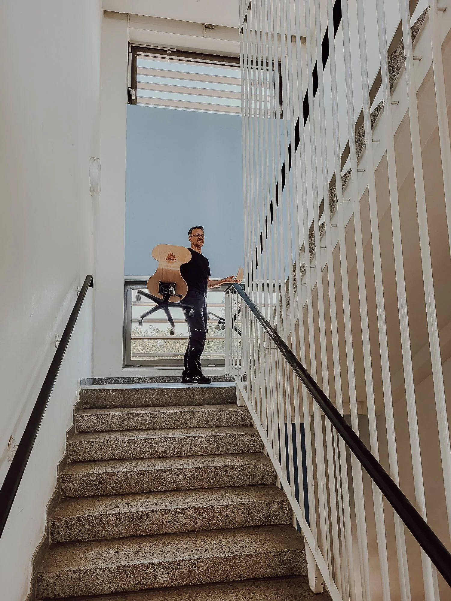 Ein Mann hält eine Gitarre in der Hand, während er auf einer Treppe in einem modernen Gebäude steht, mit weißen Wänden und einer großen Fensterfläche im Hintergrund.