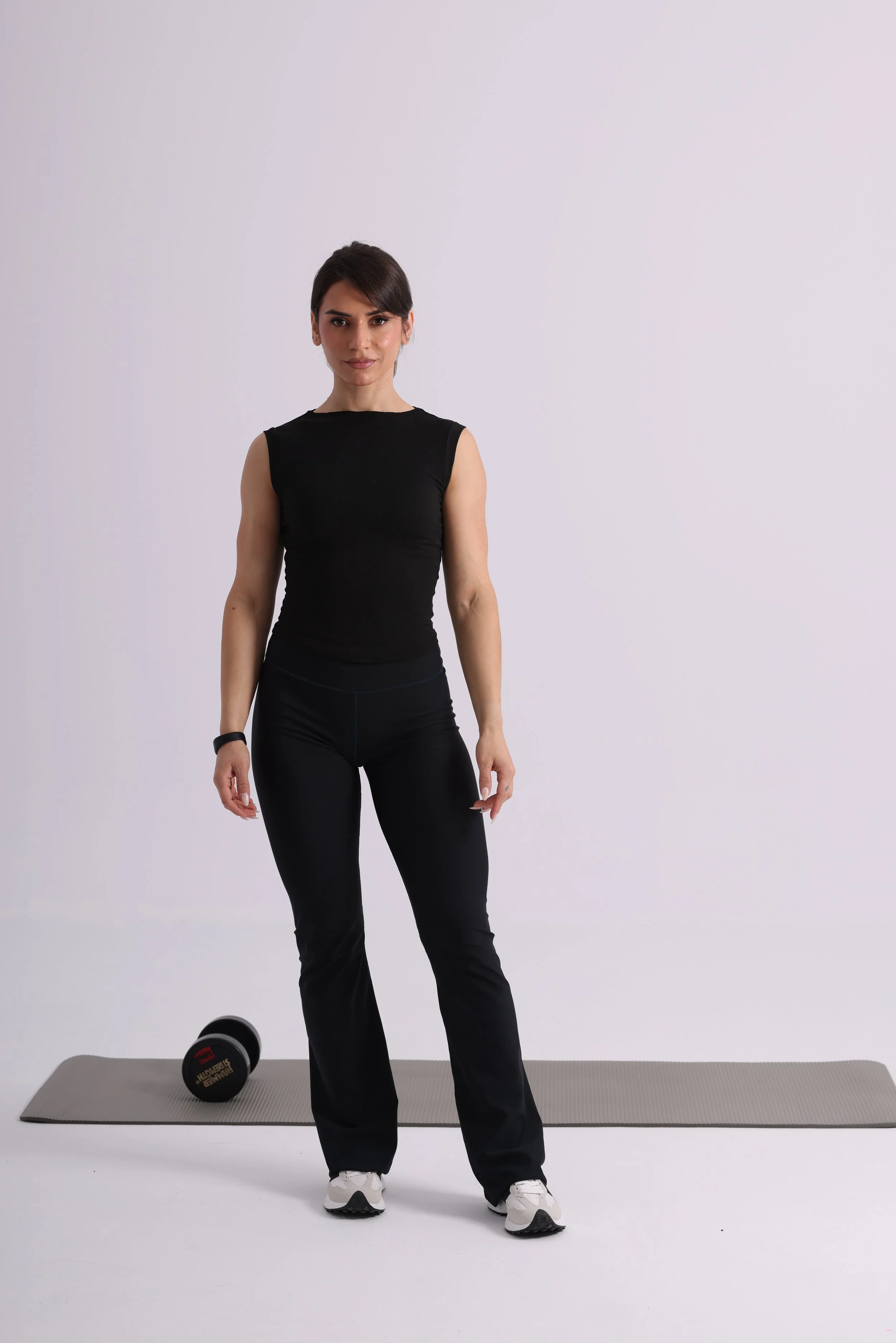 A woman standing on a yoga mat with a dumbbell lying next to it, dressed in black workout clothes and white athletic shoes, against a plain white background.