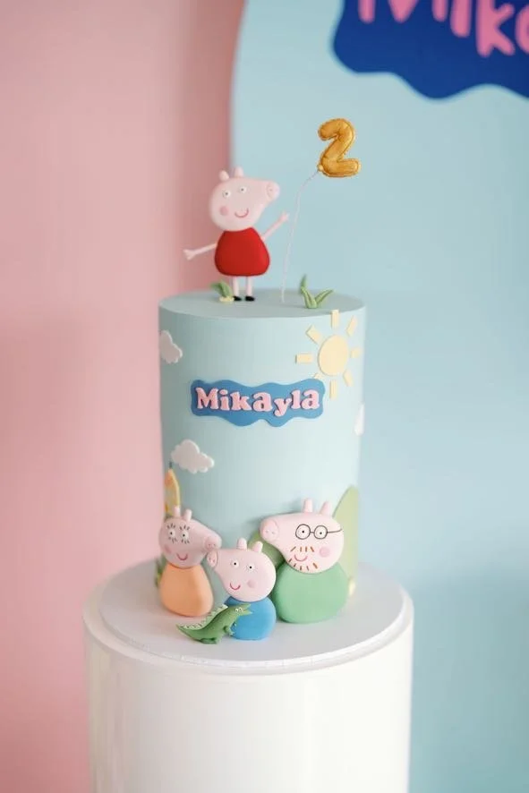 Decorative birthday cake featuring Peppa Pig characters, with a small Peppa Pig figurine holding a gold '2' balloon on top, pink, blue, and pastel colors, and a personalized name "Mikayla" in colorful lettering.