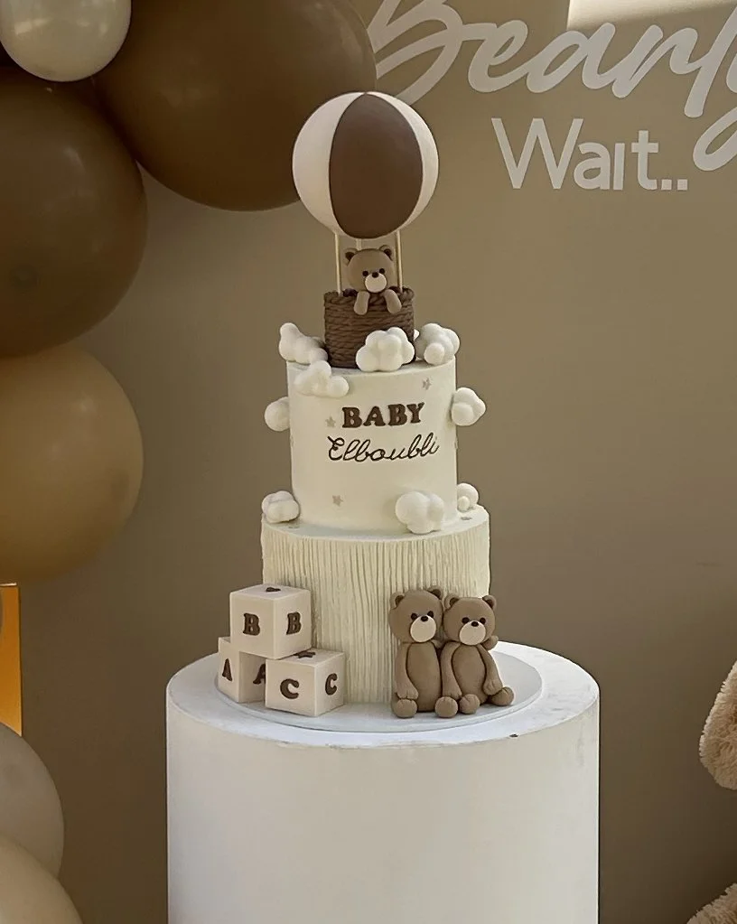 Decorative baby shower cake with teddy bear figurines, blocks, and a balloon topper, set against a beige background with balloons.