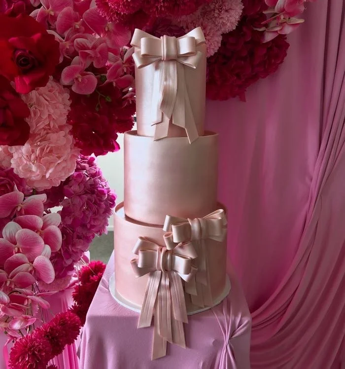 Three-tier pink cake decorated with satin bows, surrounded by pink and red flowers and pink draped fabric.