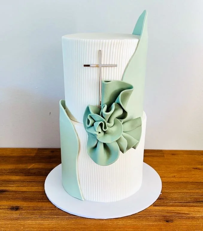 Elegant white and sage green baptism or celebration cake with a cross and ruffled fondant decoration on a white cake board.