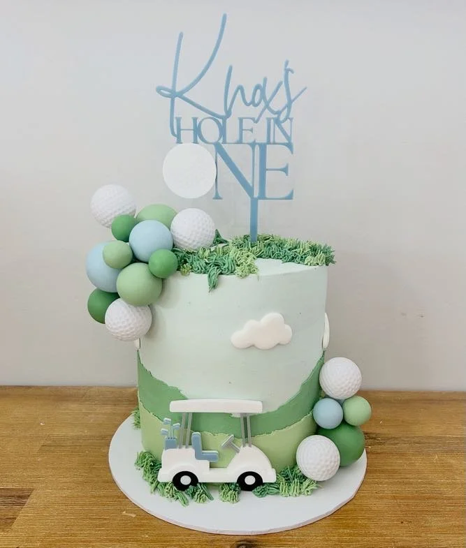 A white birthday cake decorated with green and white golf ball-themed accents, a small golf cart topper, and a "Knight of One" cake topper, set on a wooden table.