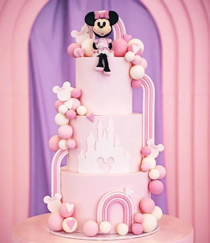 Three-tier pink birthday cake with Minnie Mouse plush toy on top, decorated with pink and white balls, Mickey Mouse silhouettes, and rainbow-shaped fondant accents in a pastel pink and purple background.