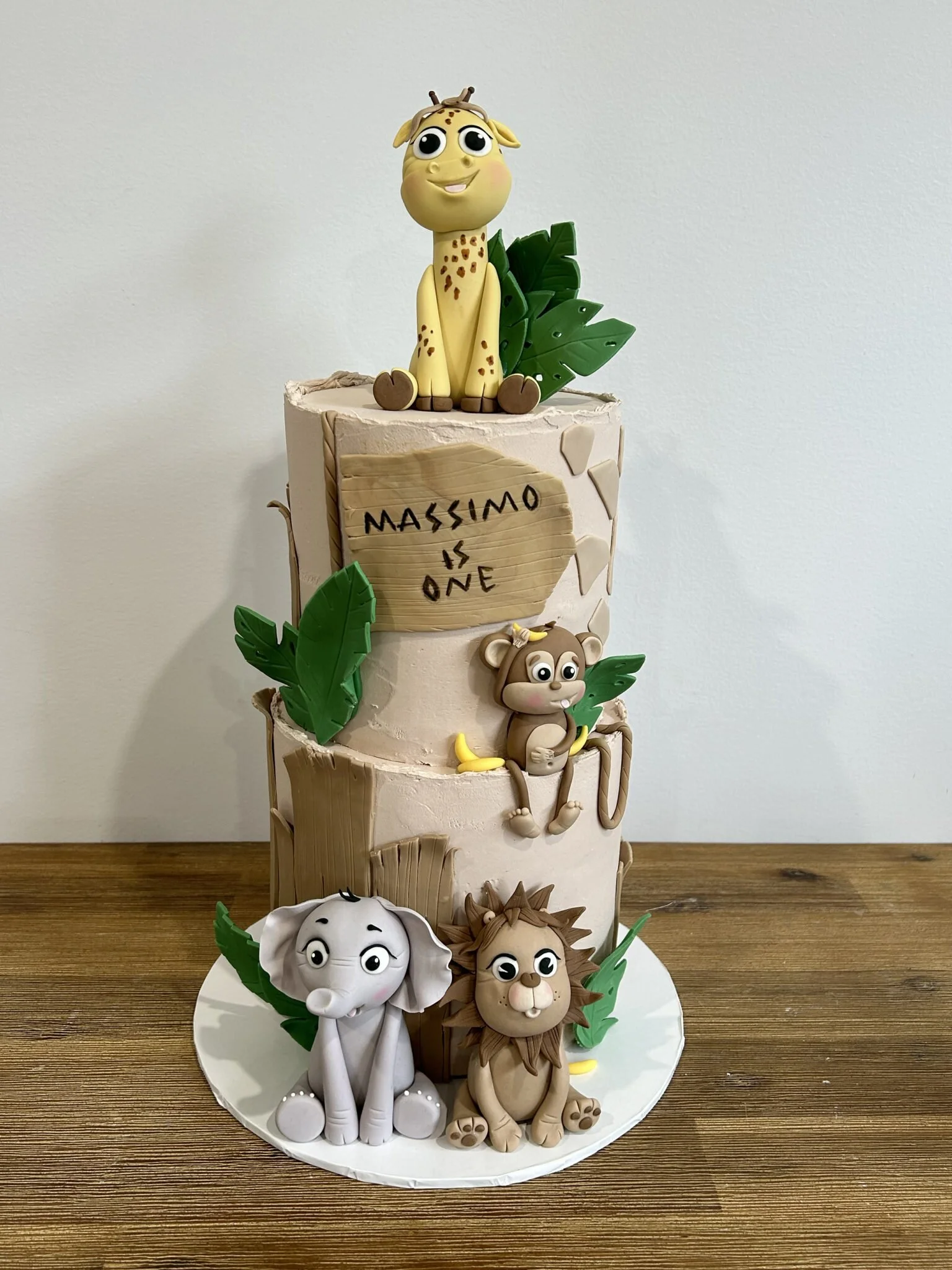 Three-tier birthday cake decorated with jungle animal figures including a giraffe, monkey, elephant, and lion, with green leaves and a wooden sign reading "Massimo is One."