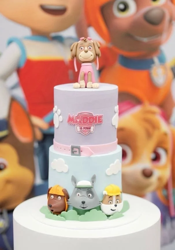 A cake decorated with characters from the animated series 'Madeline is Four,' featuring figurines of dogs and cats on top and around the cake, with a large blue and pink theme, and a background of large character balloons.